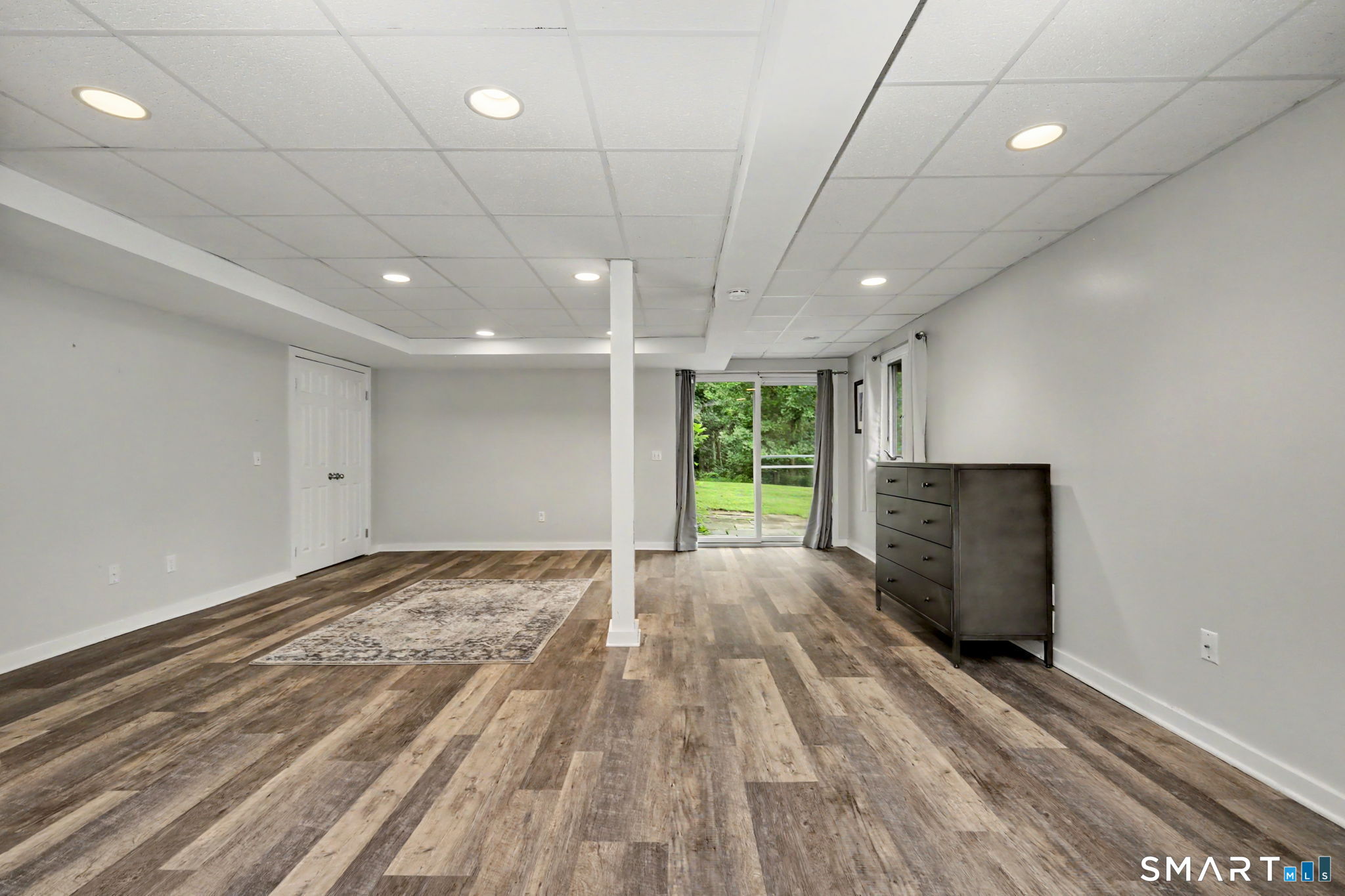Finished lower-level room with wood-look floors, a central support column, and sliding doors to the yard.
