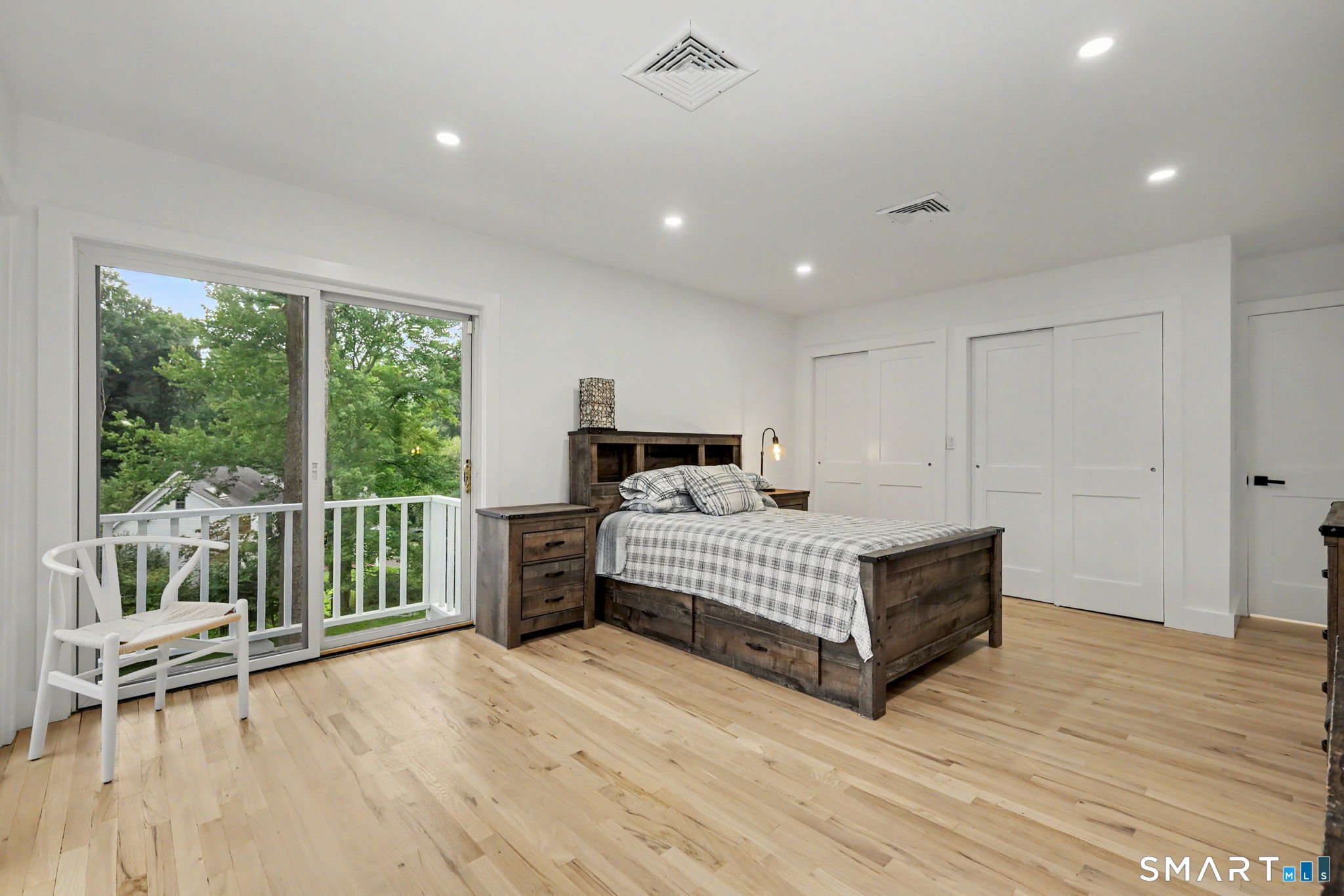 Bedroom with a dark wood bed, light hardwood floors, and sliding doors to a tree-lined balcony.
