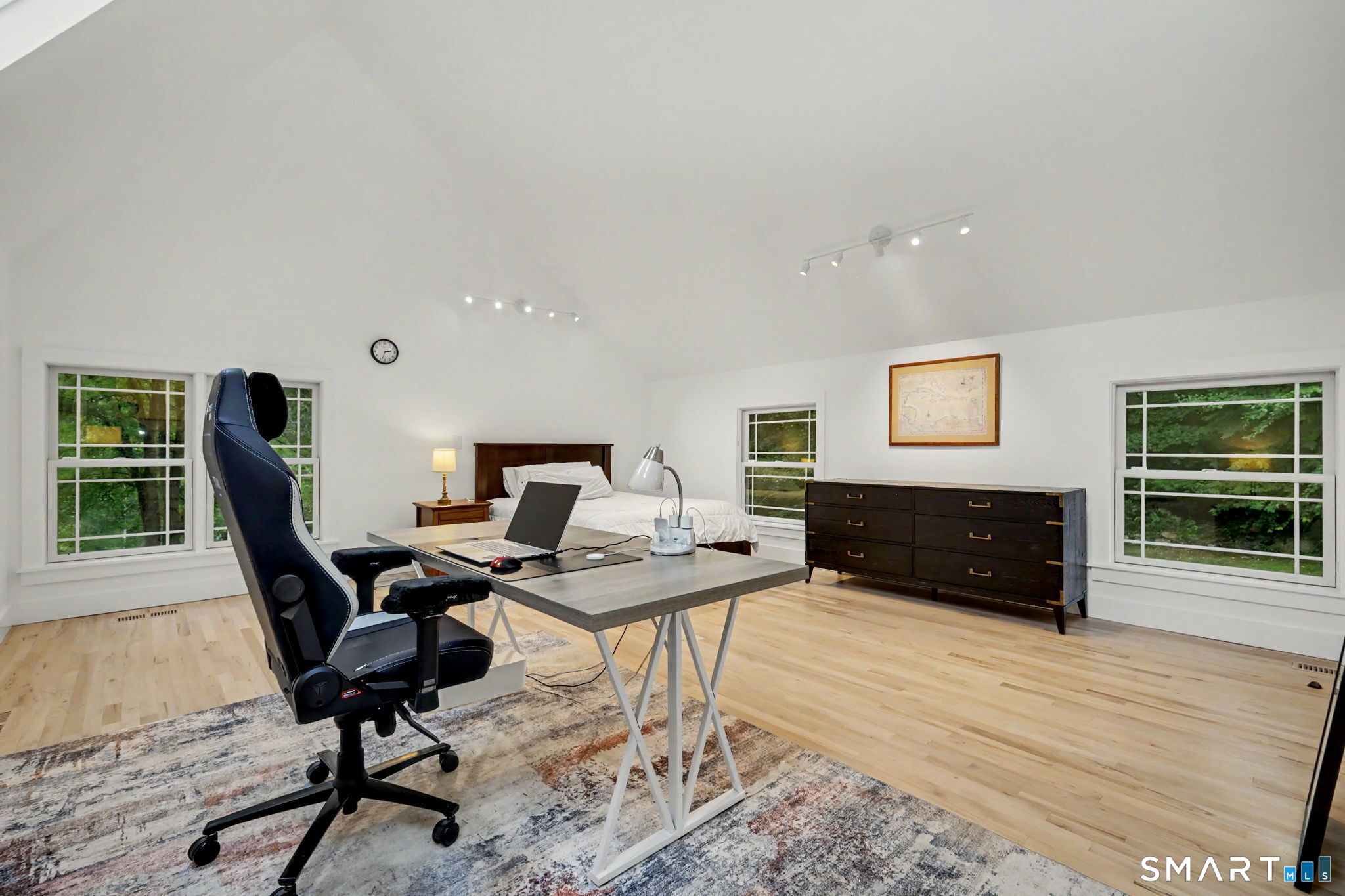 Bedroom with a vaulted ceiling, desk and laptop, black office chair, and windows overlooking greenery.