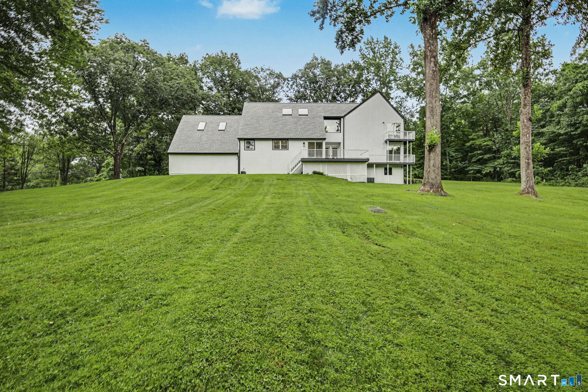 White multi-level house with decks overlooking a broad lawn and mature trees.