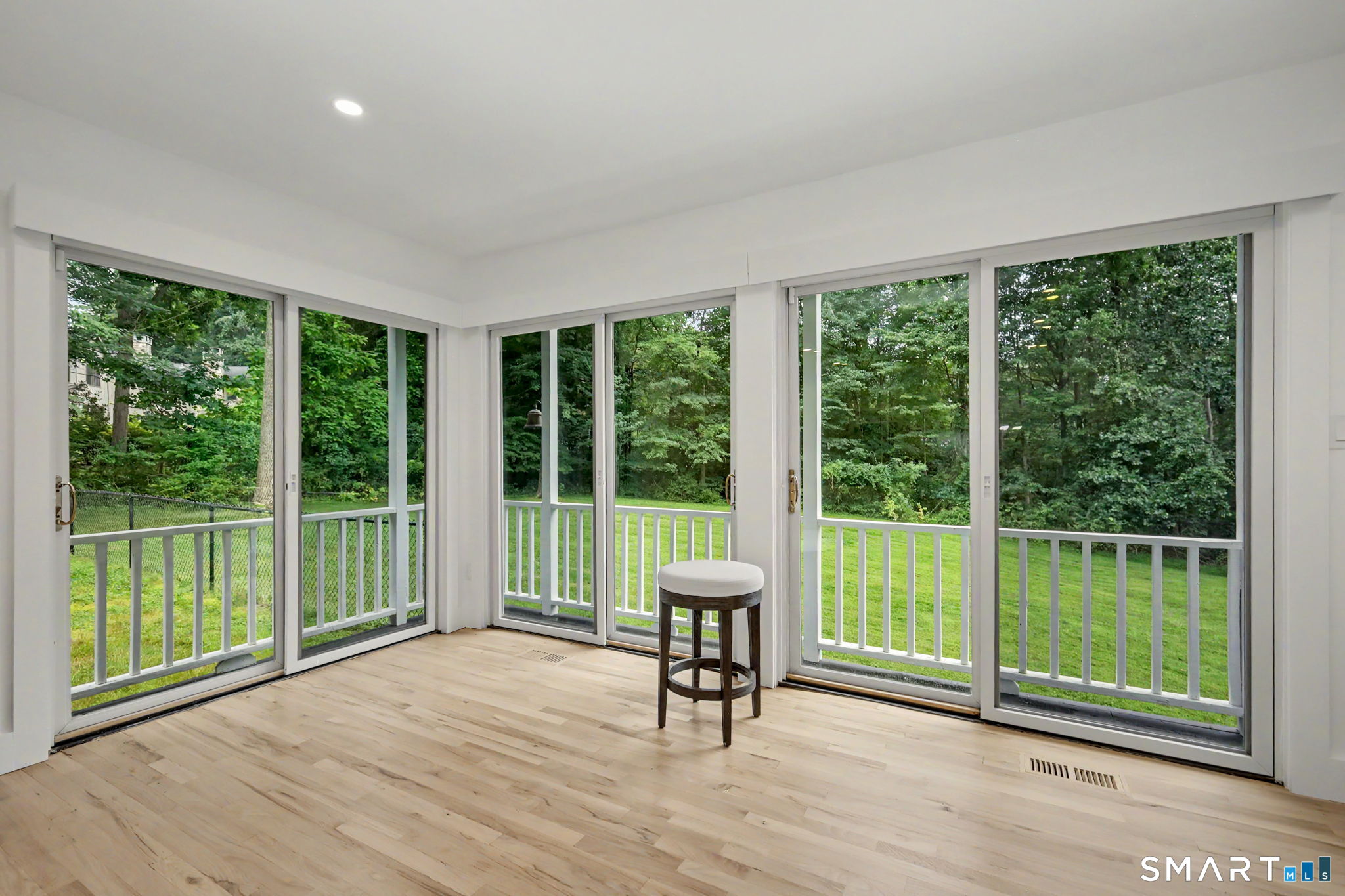 Bright sunroom with pale wood floors, a single stool, and wide windows overlooking a wooded lawn.