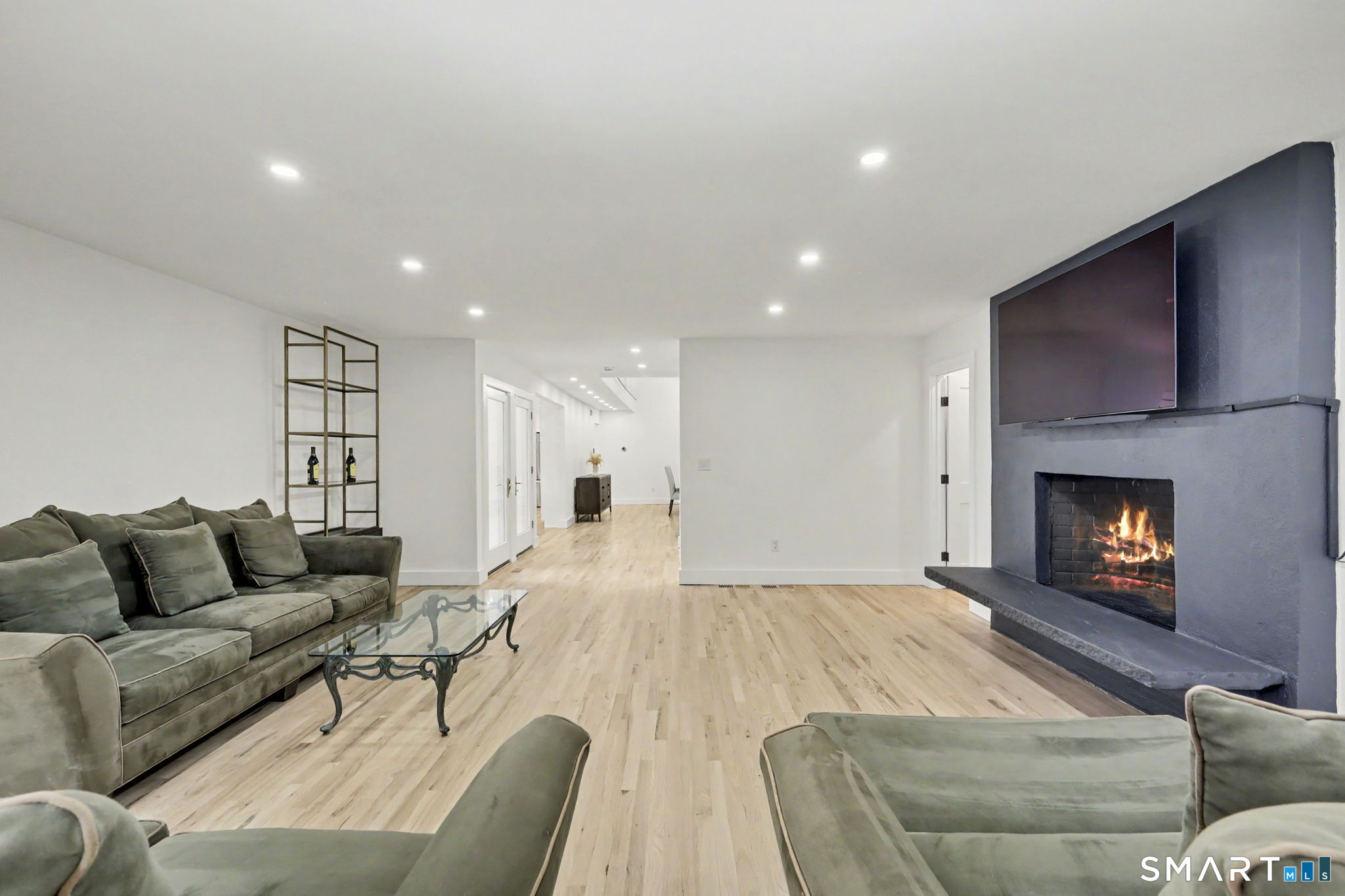 Living room with pale hardwood floors, gray-green sofas, and a lit fireplace beneath a mounted television.