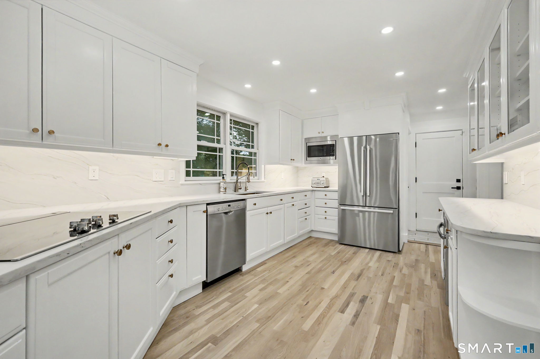 White kitchen with stainless steel appliances, a window above the sink, and light wood floors.