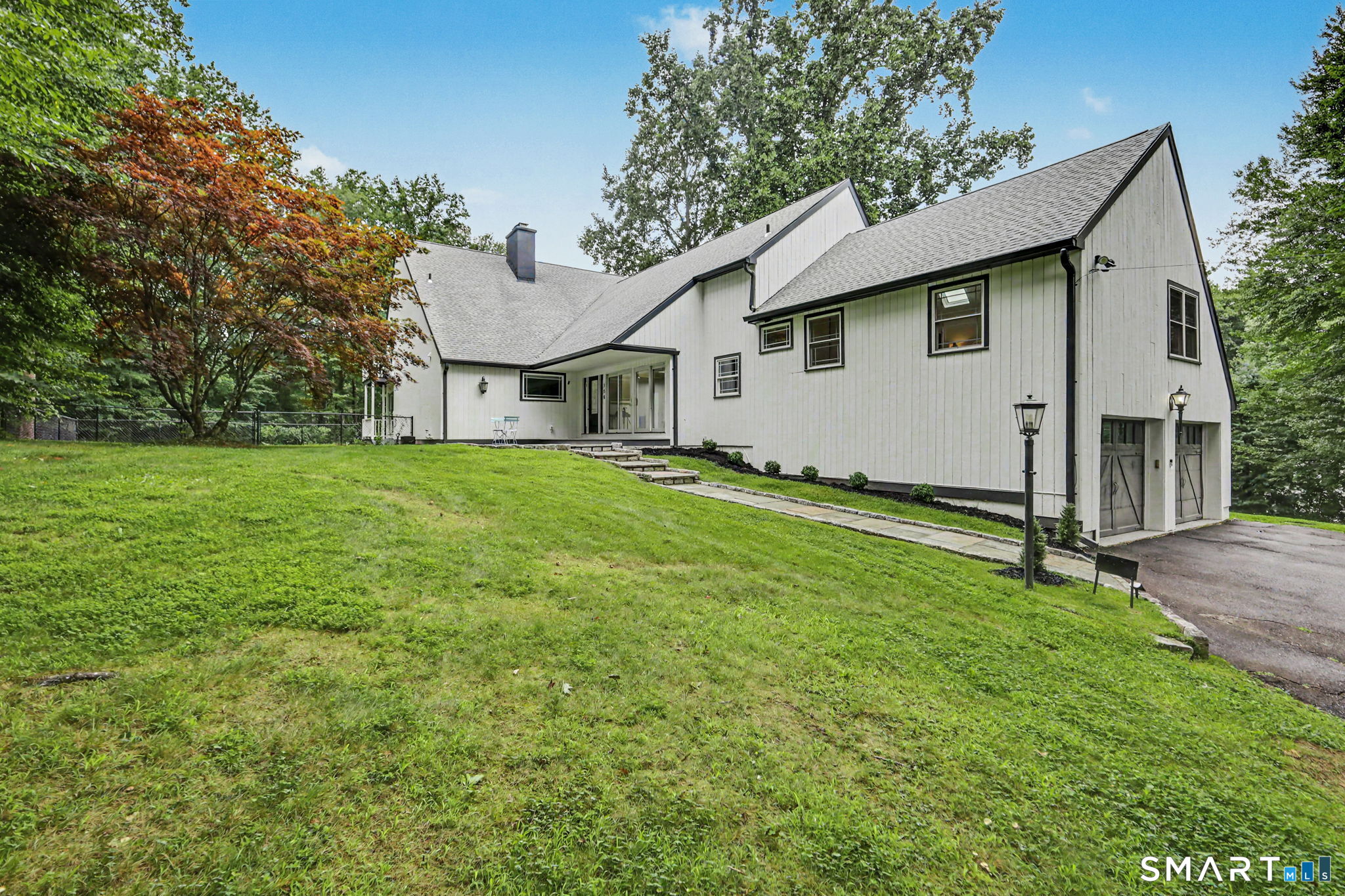 White gabled house with garage, stone walkway, and sloping lawn among mature trees.