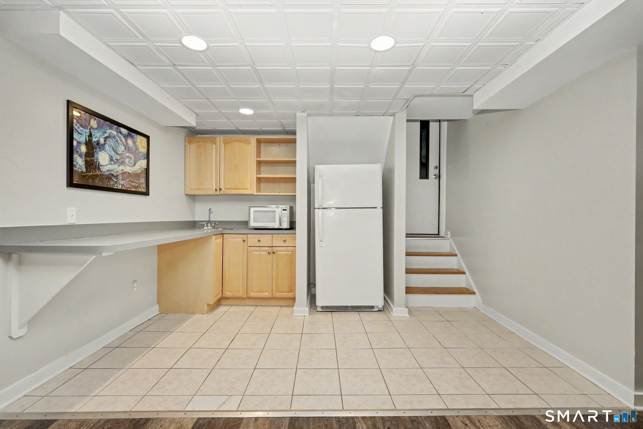 Compact kitchen with light wood cabinets, white refrigerator, tiled floor, and steps to a door.