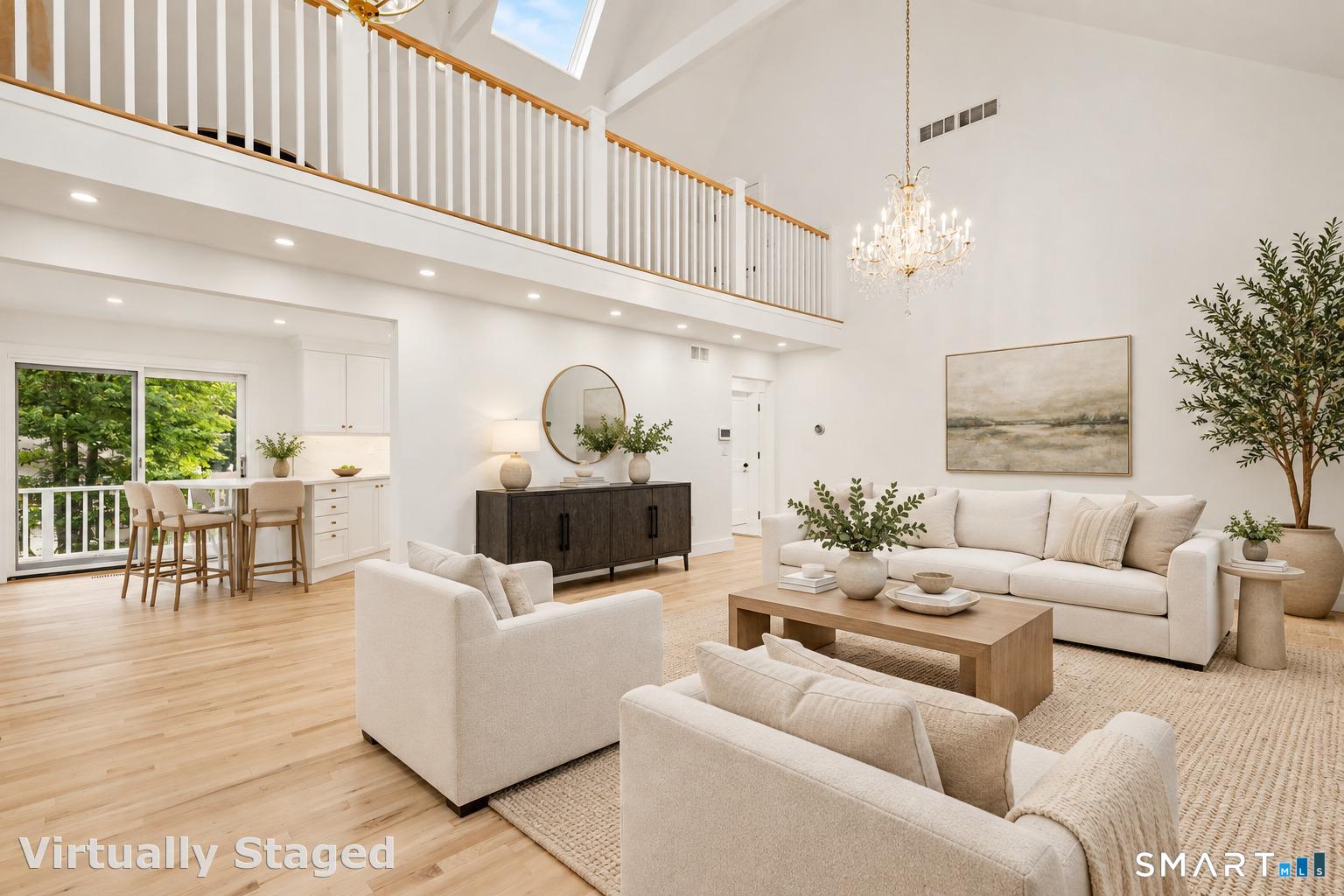 Double-height living room with a mezzanine balcony, chandelier, white seating, and an adjoining dining area.