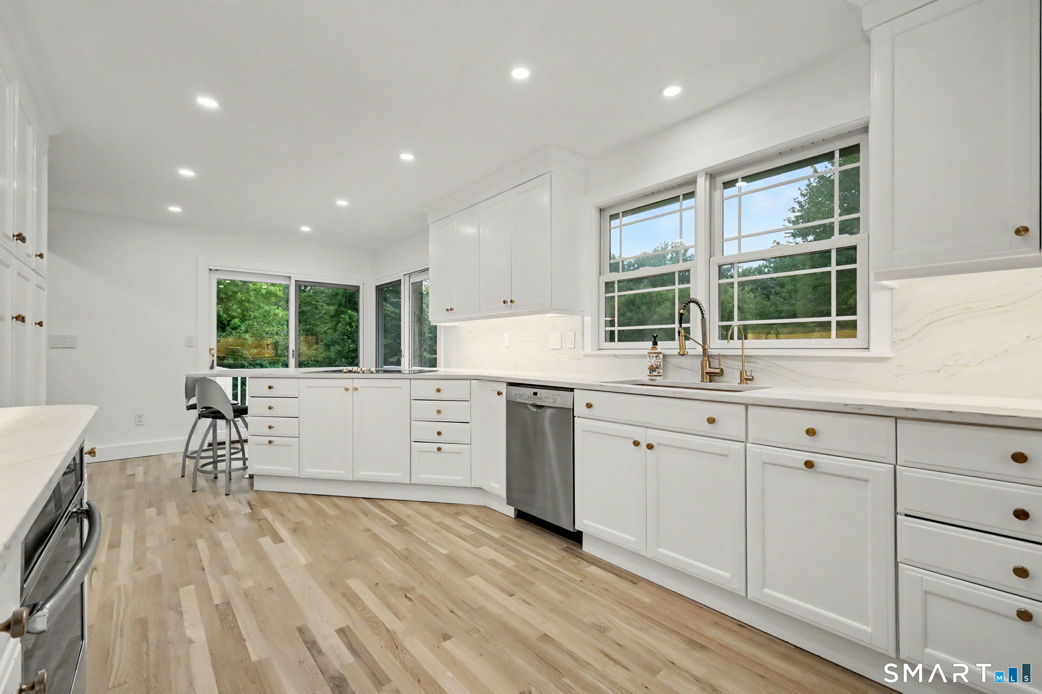 Bright white kitchen with brass hardware, light wood floors, stainless steel appliances, and large windows.