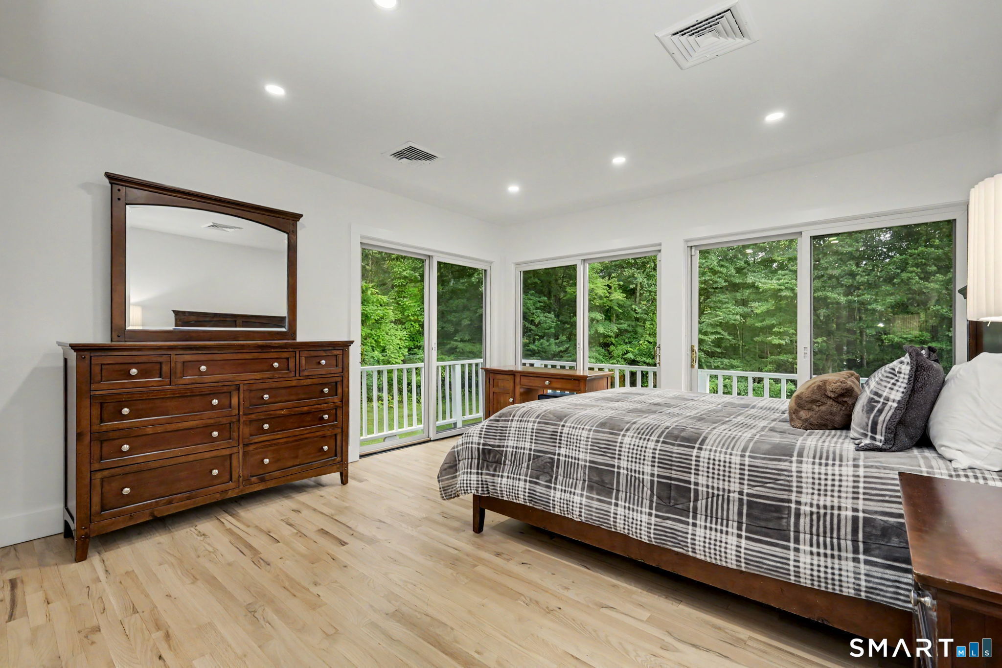 Bedroom with gray plaid bedding, a dark wood dresser, and large windows overlooking leafy woods.