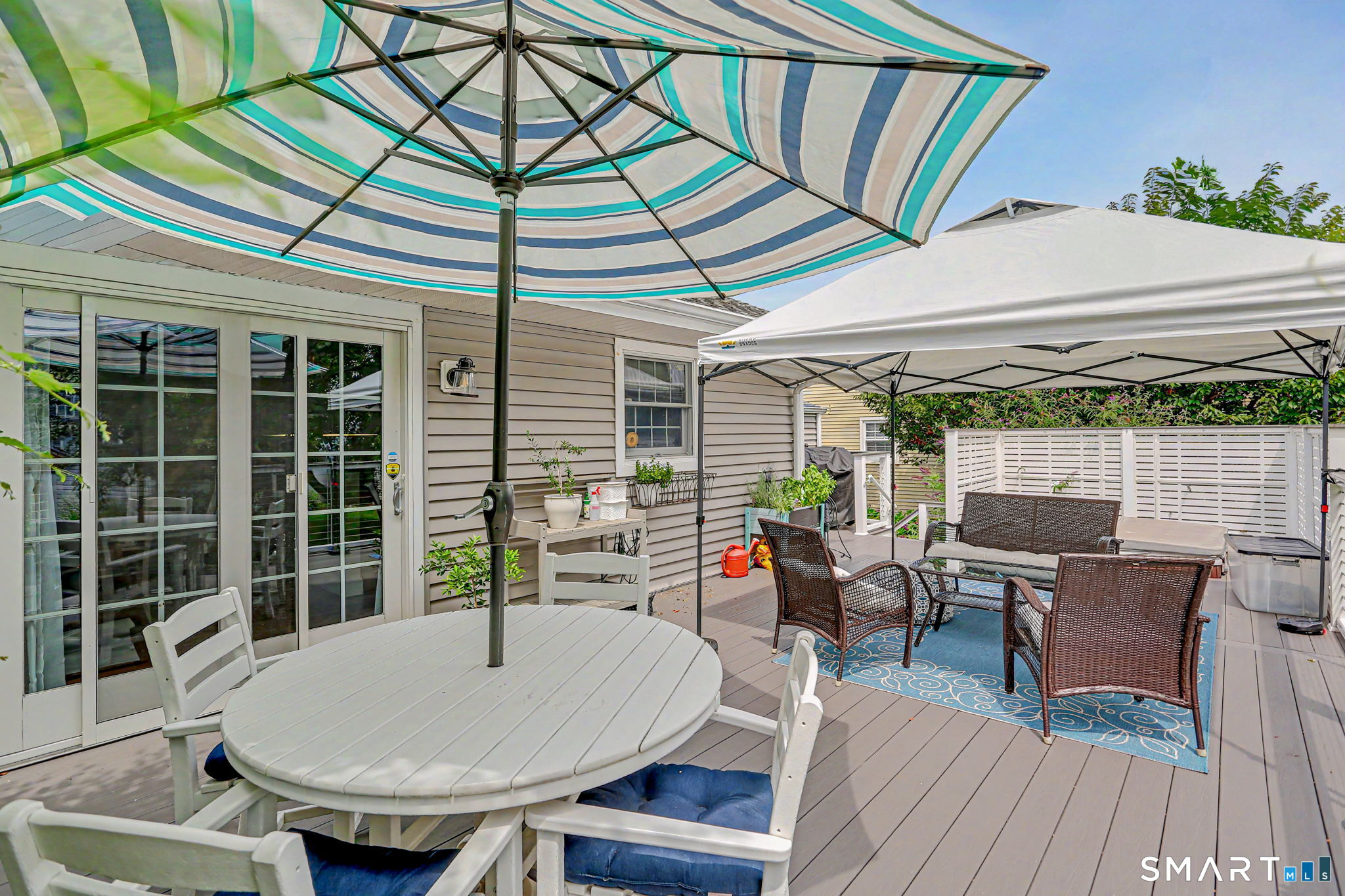 Backyard deck with striped umbrella, round dining table, and wicker seating under a white canopy.