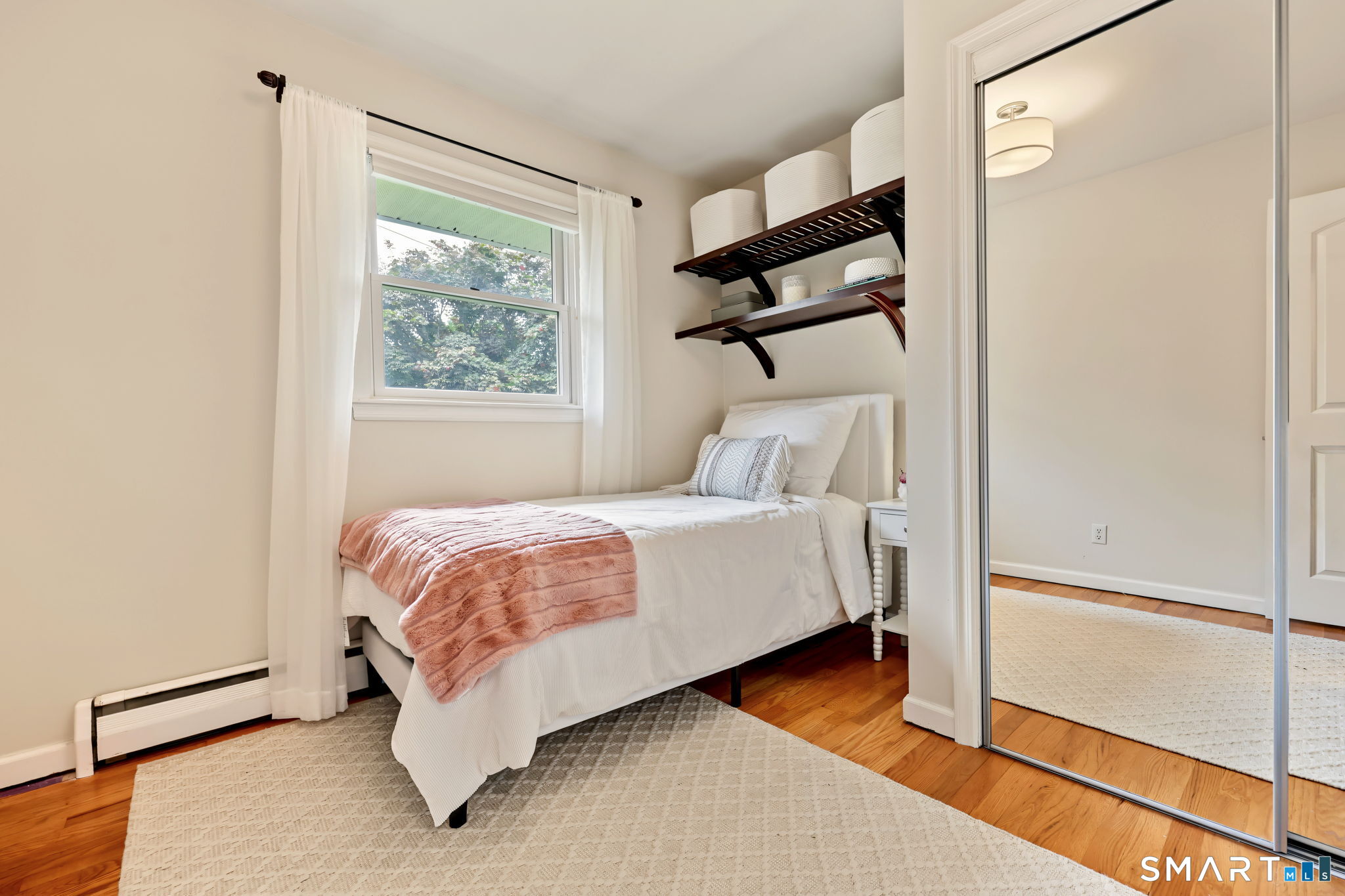 Bedroom with a white bed, blush throw, window, and mirrored closet doors.