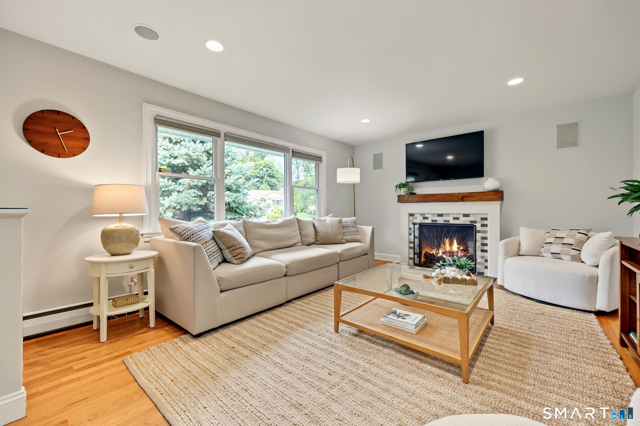 Furnished living room with a lit fireplace, large windows, and hardwood floors.