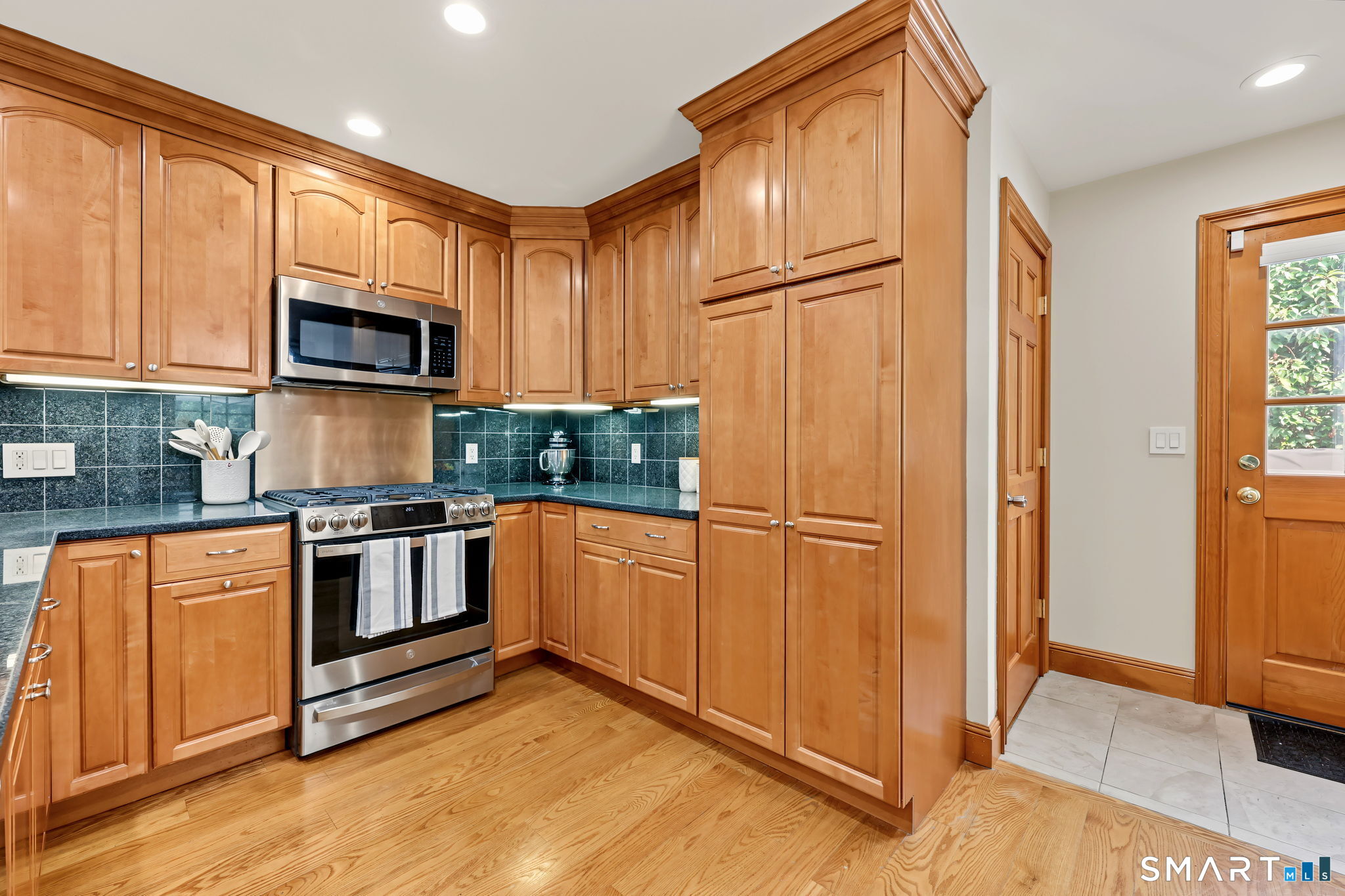 Kitchen with honey-colored wood cabinets, dark tile backsplash, stainless steel range, and hardwood floors.