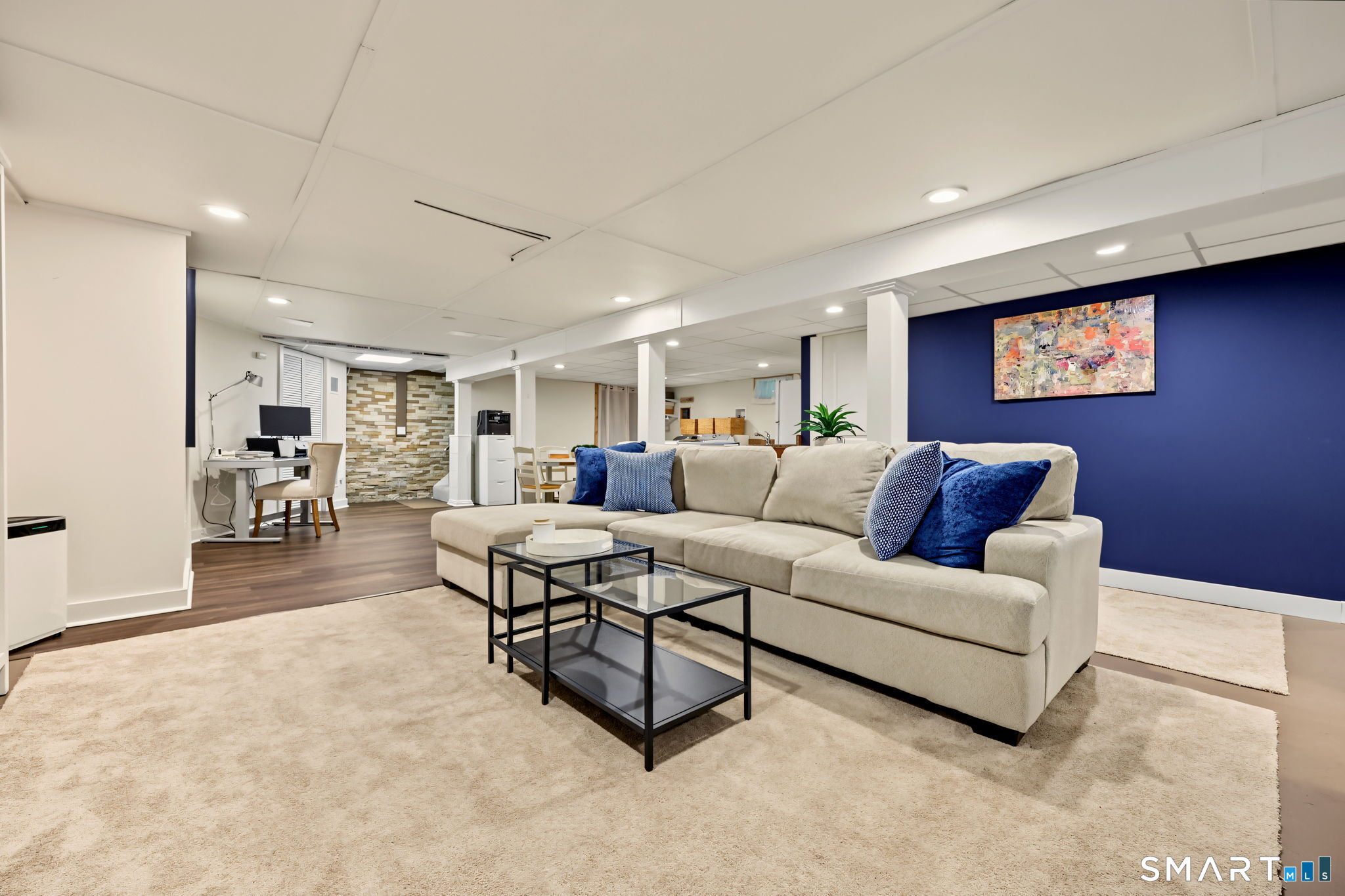 Basement living room with a cream sectional, blue accent wall, and adjoining desk area.