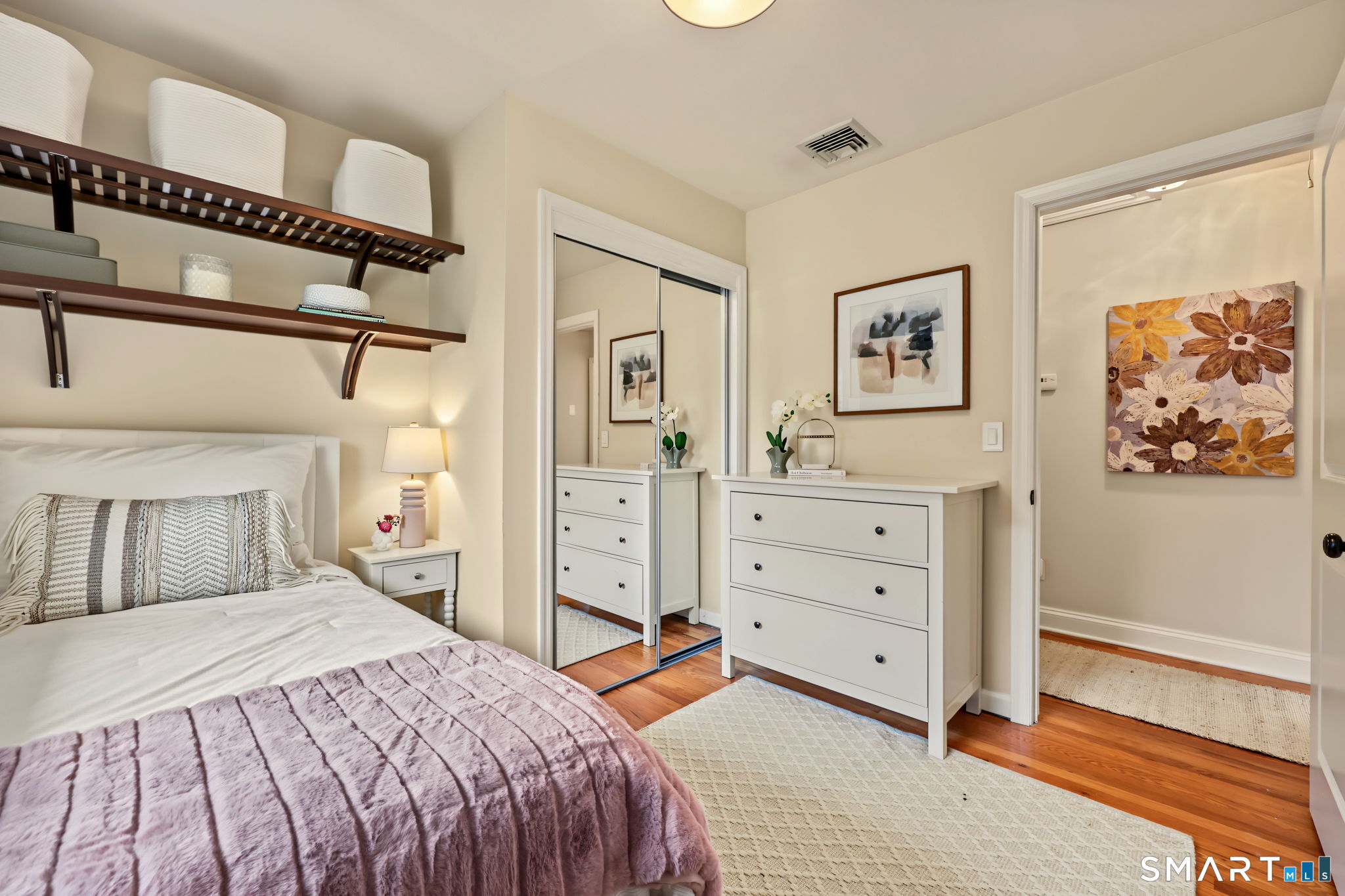 Staged bedroom with a mauve quilt, floating shelves, mirrored closet doors, and a white dresser.