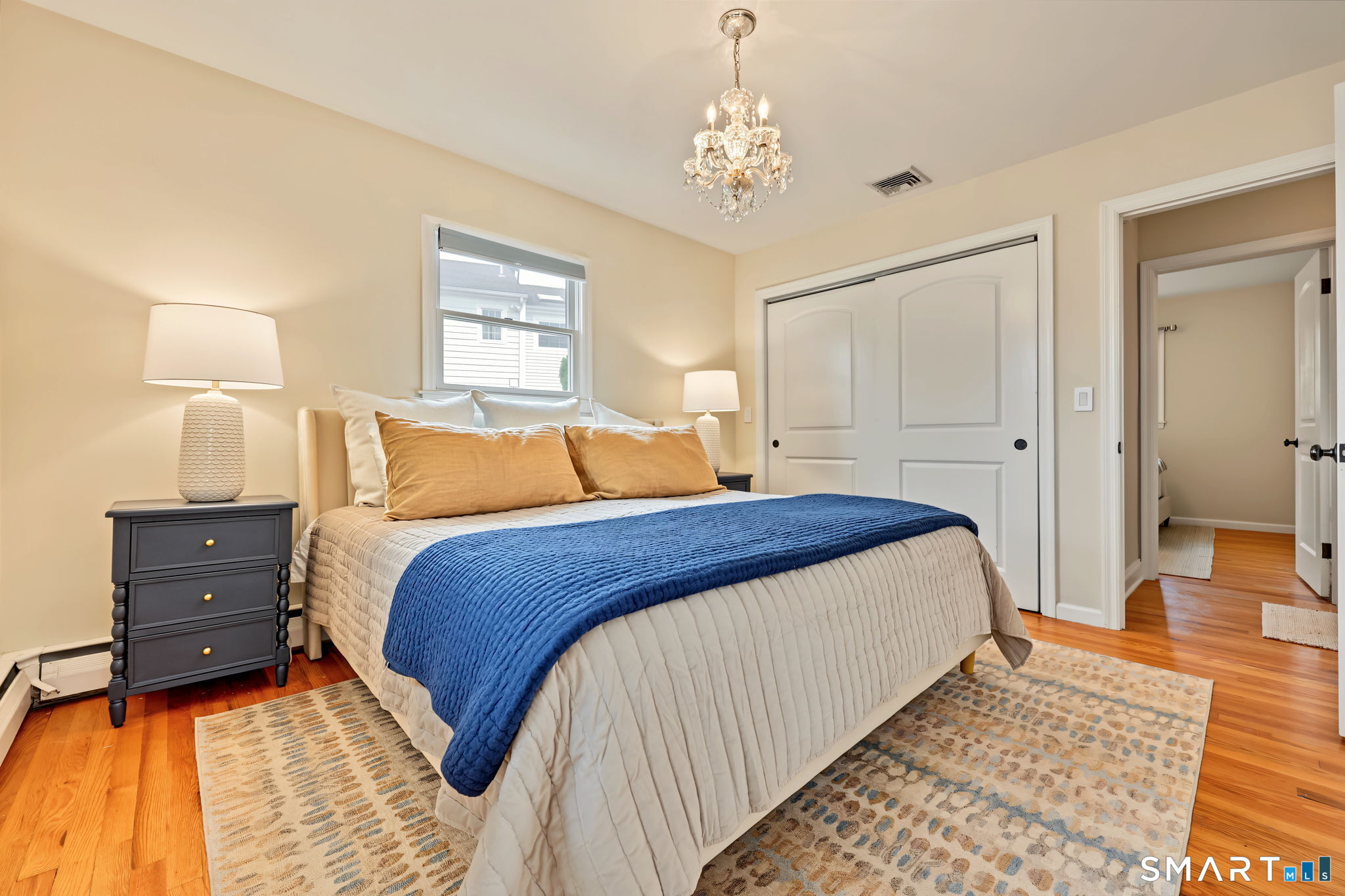 Furnished bedroom with a blue bedspread, crystal chandelier, hardwood floors, and patterned rug.