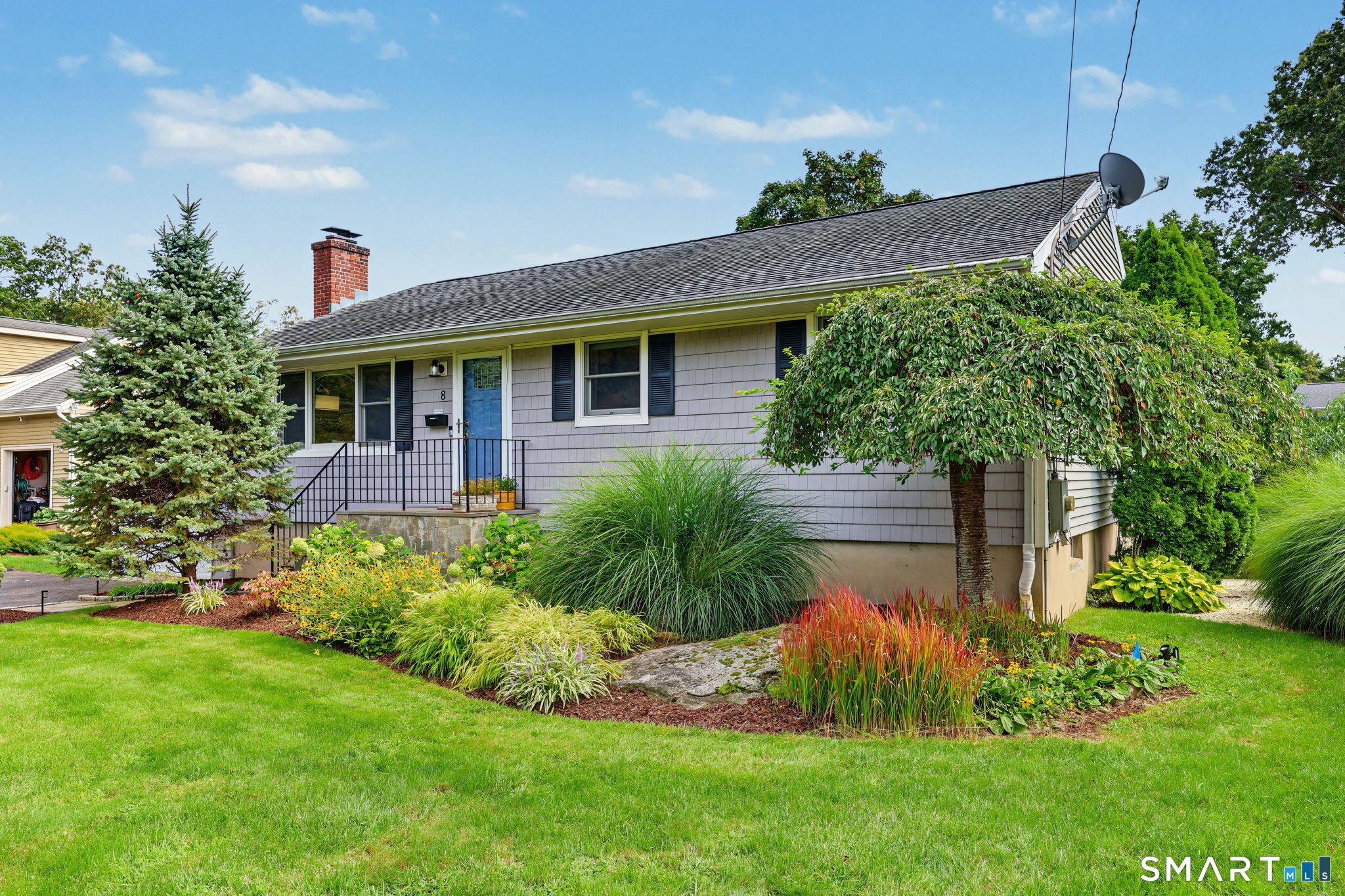 Single-story gray house with a blue front door, brick chimney, and colorful landscaped garden beds.