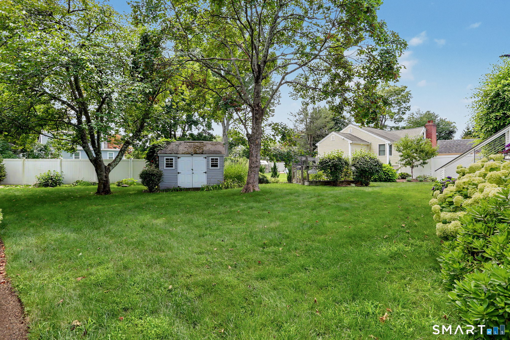 Grassy backyard with mature trees, a gray storage shed, white fencing, and shrubs.