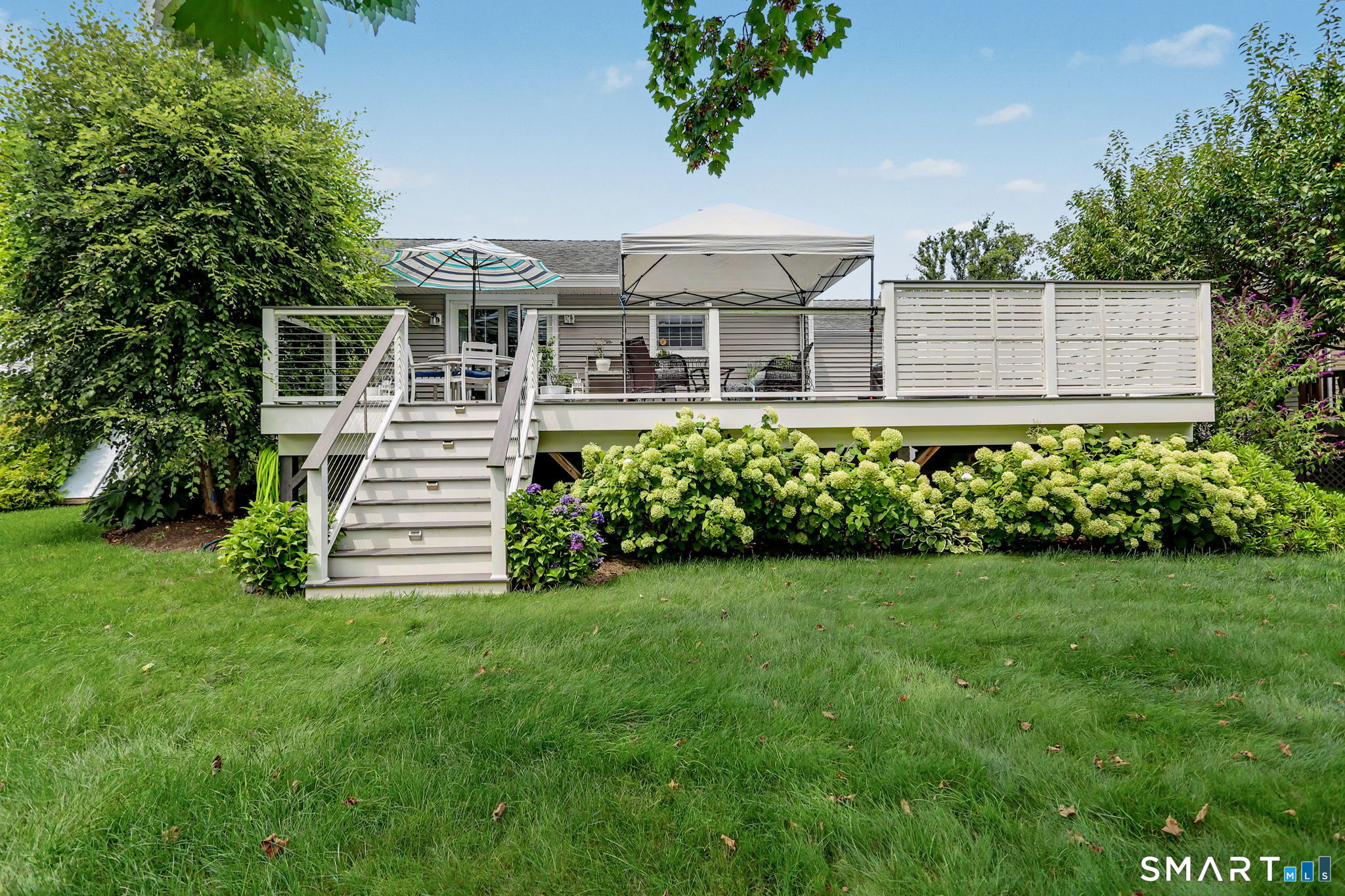 House rear deck with white railings and stairs, patio seating under a canopy, and flowering shrubs.