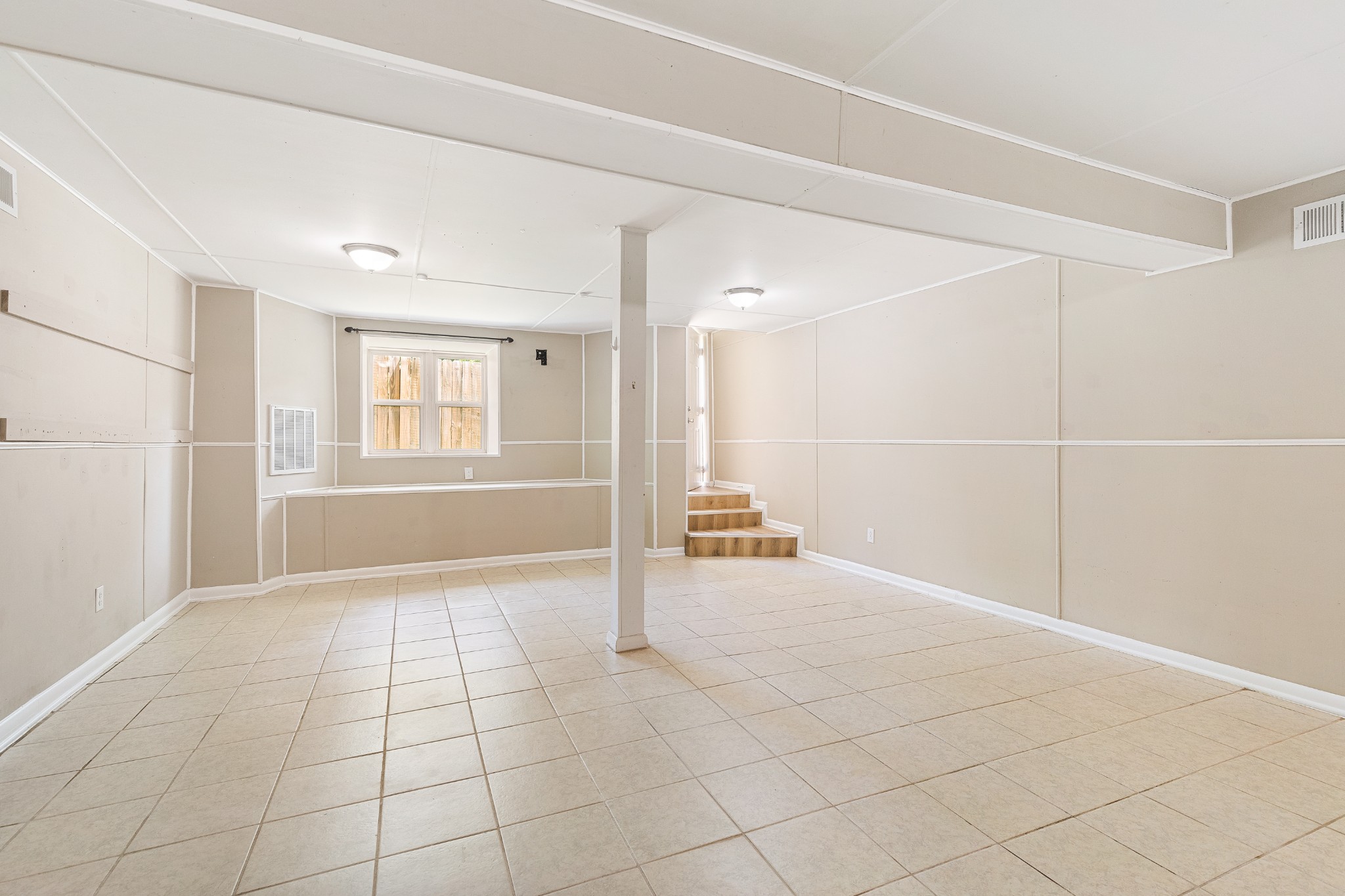 Vacant lower-level room with tiled floors, a central support post, and steps to a door.