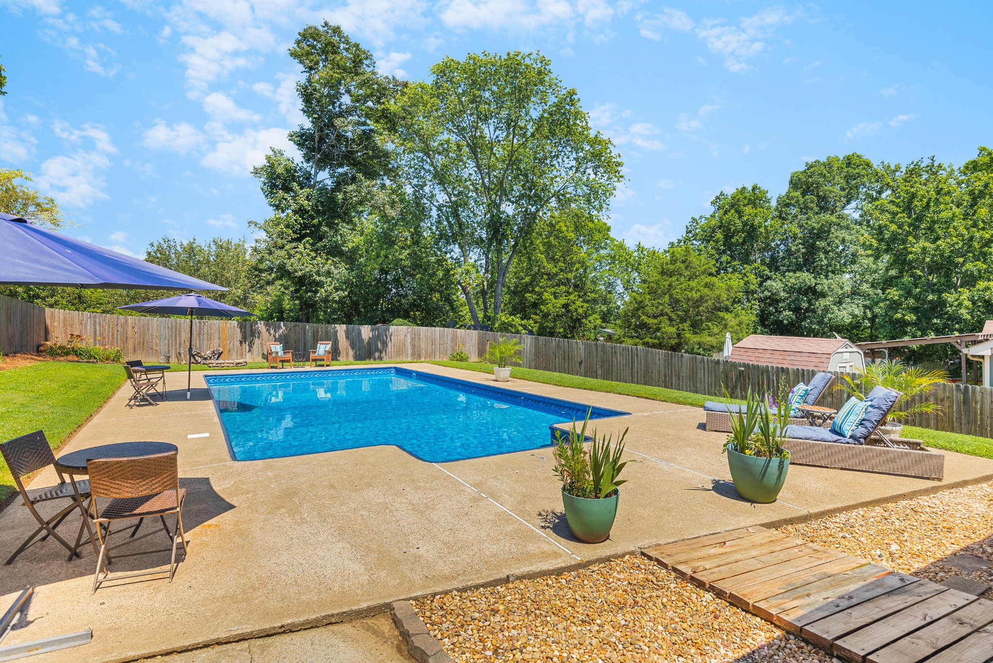 Backyard swimming pool with a fenced patio, lounge chairs, umbrellas, and mature trees.