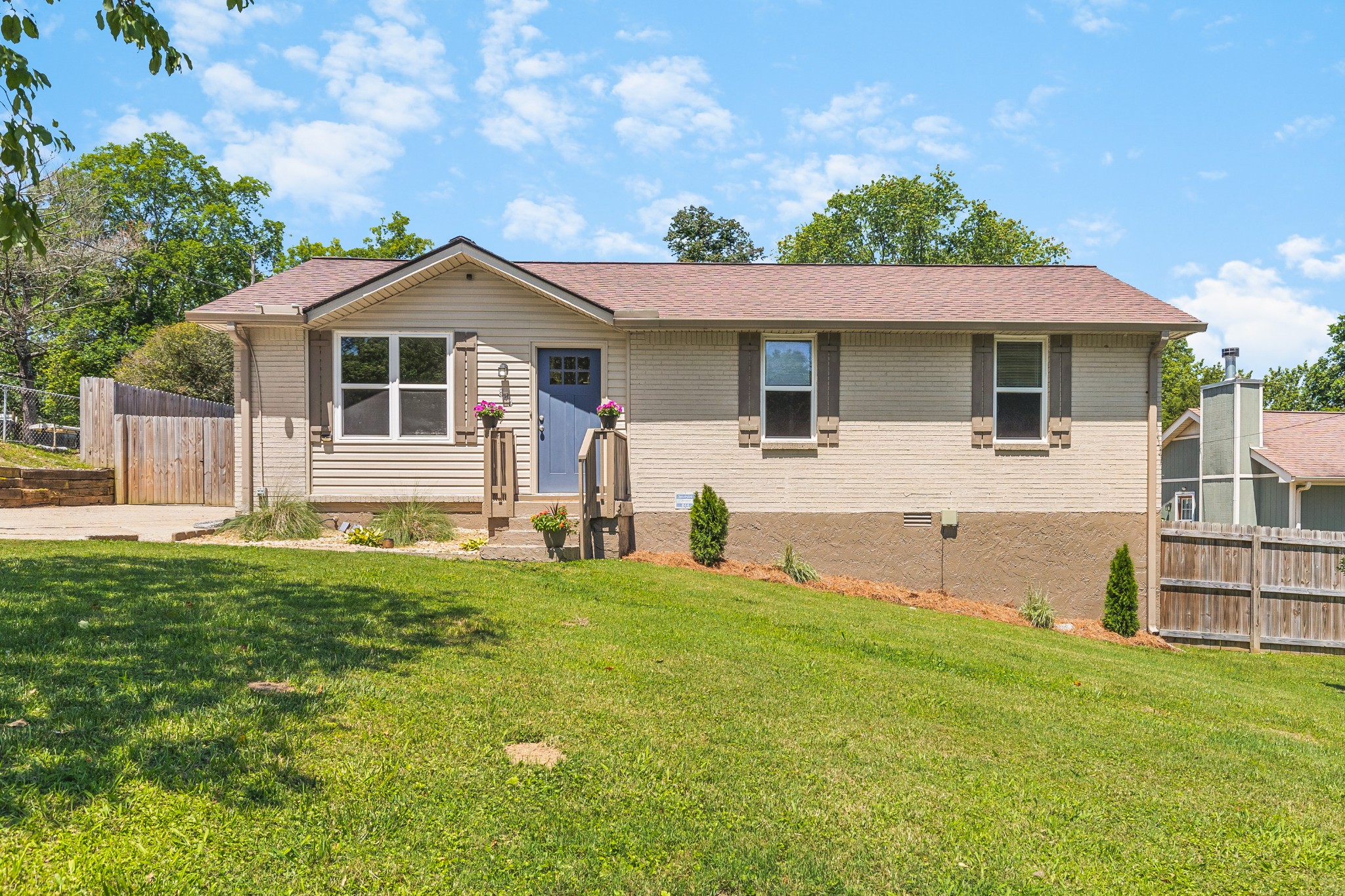 Single-story beige brick home with a blue front door, flower pots, and a broad lawn.