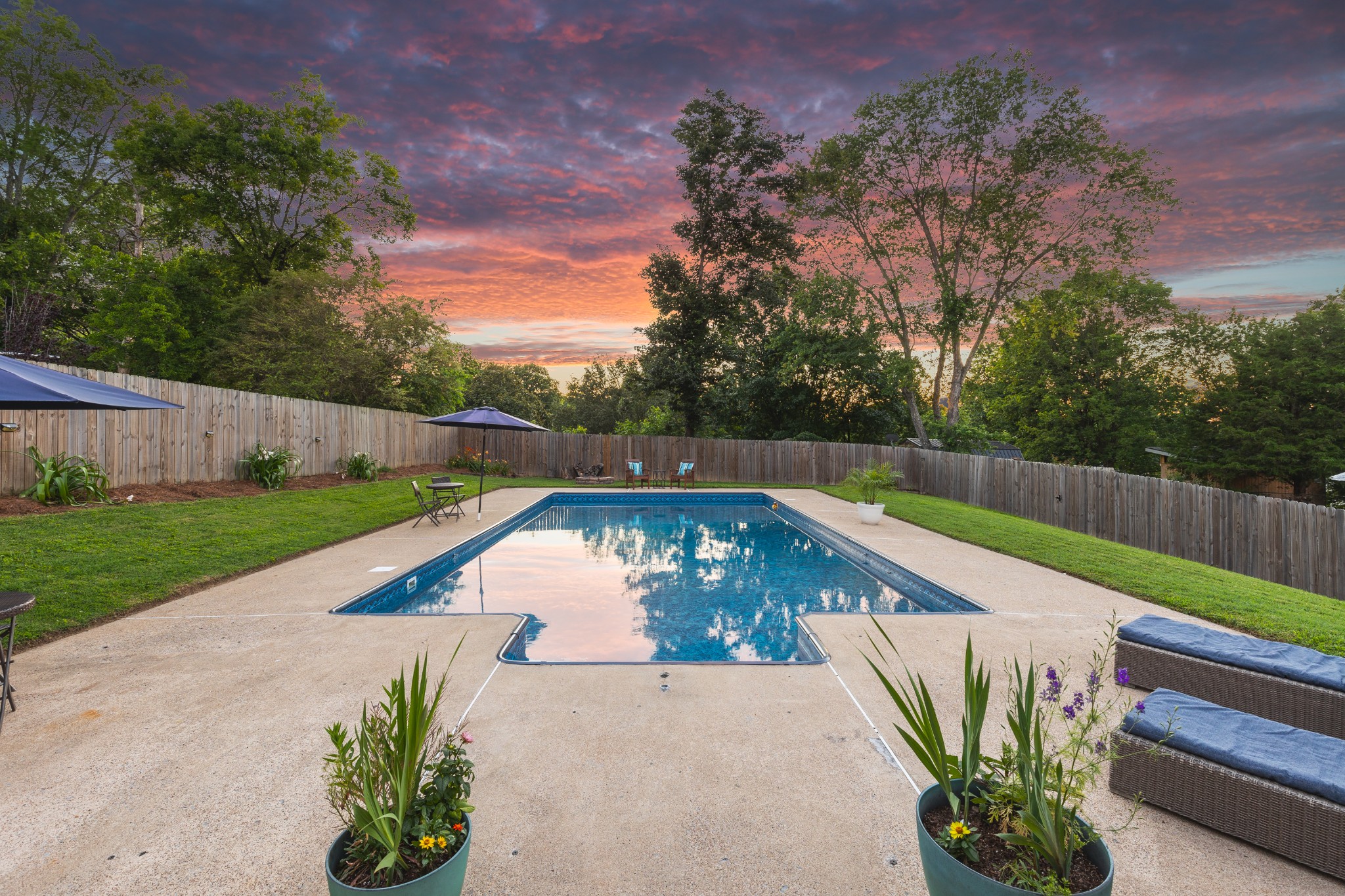 Backyard swimming pool with a wide concrete deck, patio seating, and colorful sunset sky.