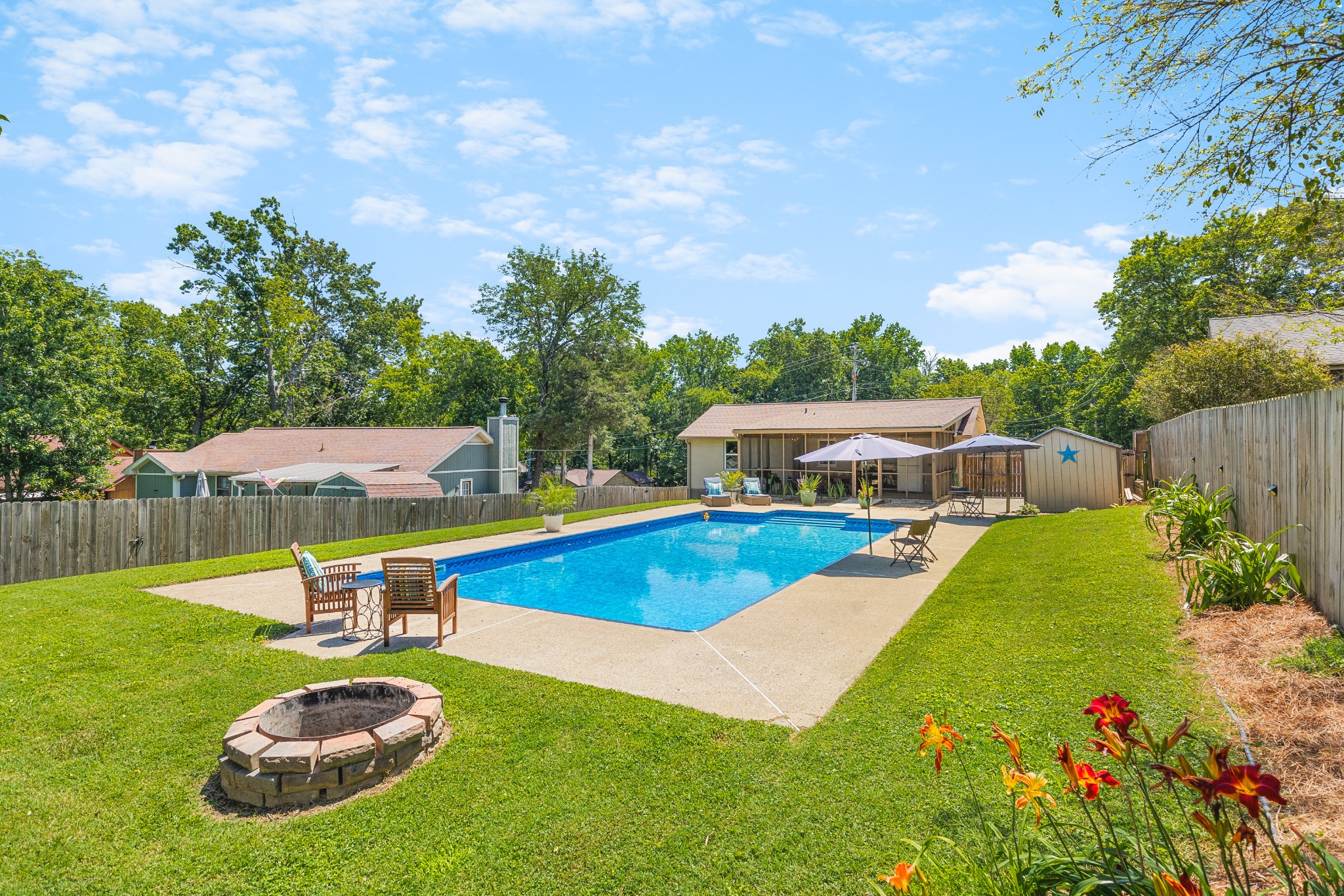 Backyard pool with patio umbrellas, seating, a stone fire pit, and colorful flowers.