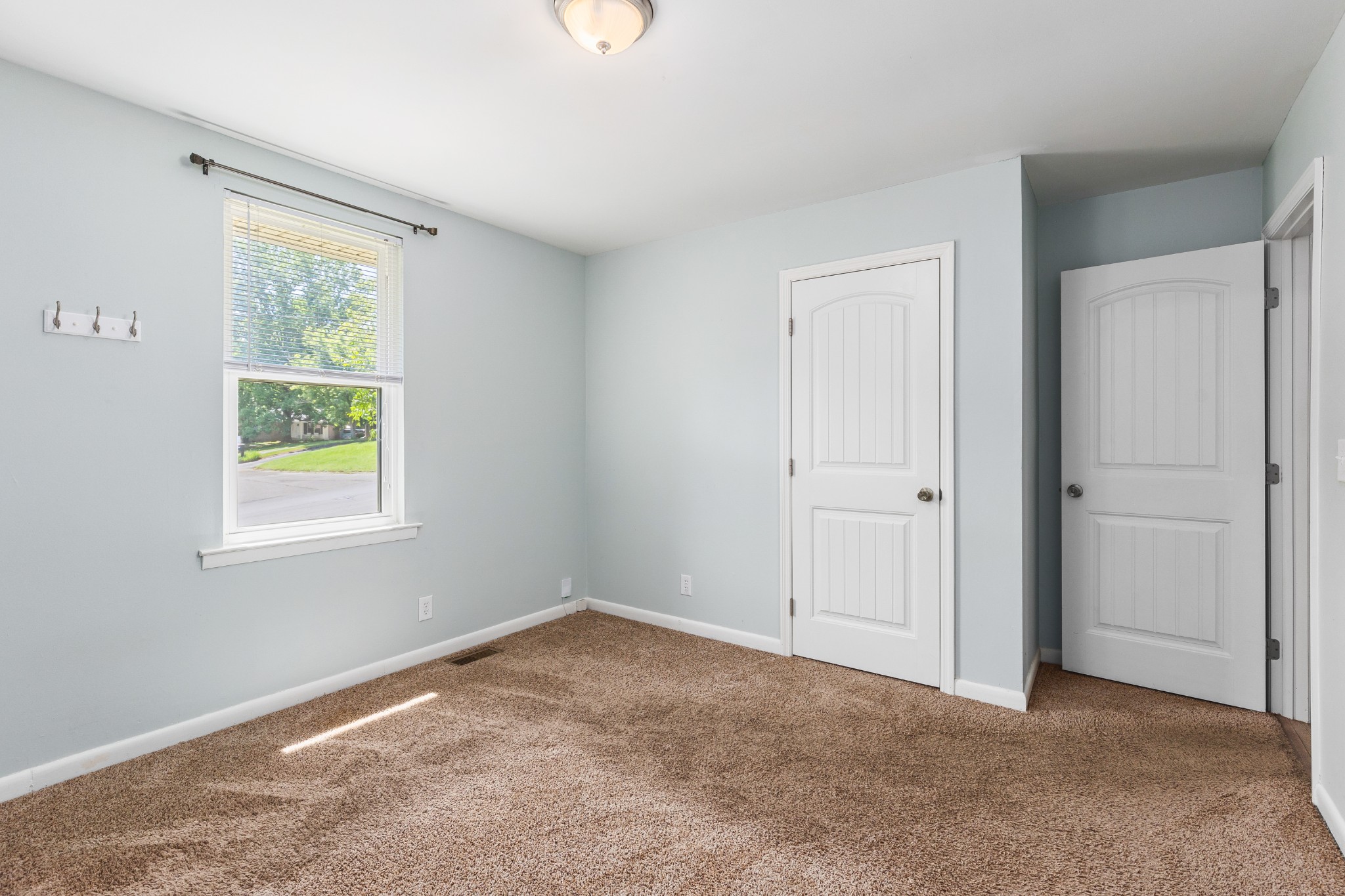Empty carpeted room with pale walls, a window with blinds, and two white paneled doors.