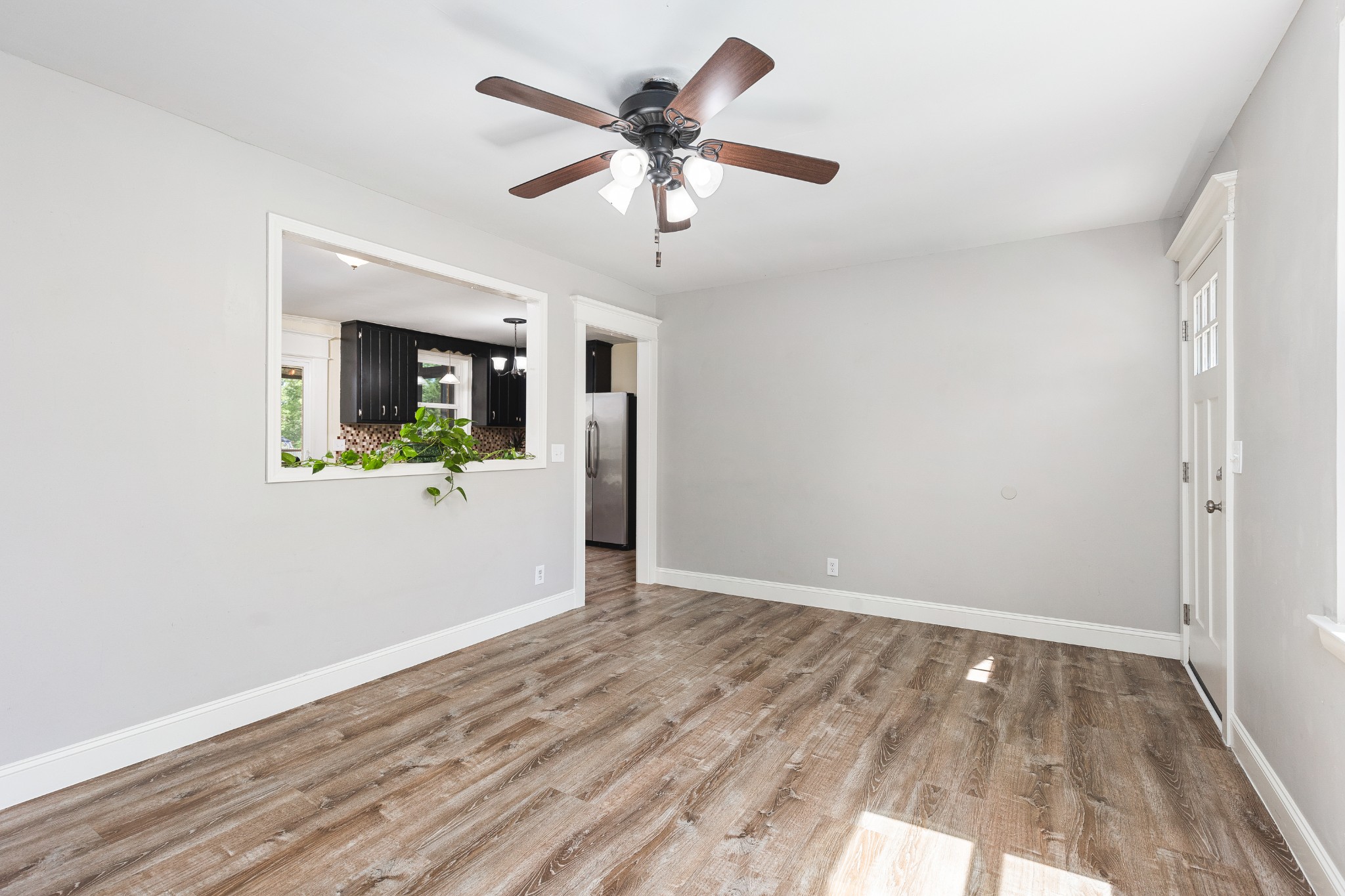Empty dining area with wood-look floors, ceiling fan, and pass-through to a dark-cabinet kitchen.