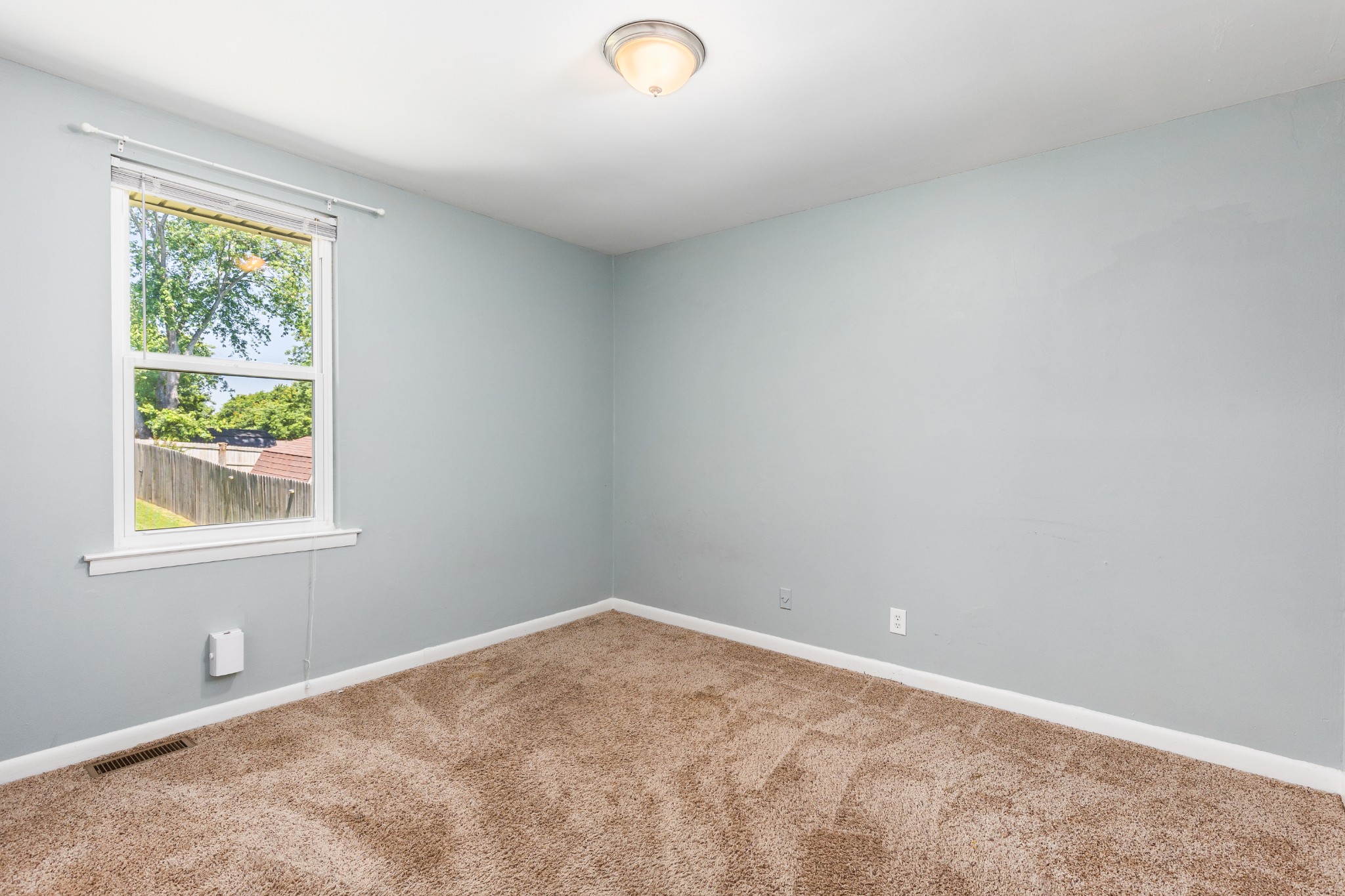 Empty carpeted room with pale blue-gray walls and a window overlooking a fenced yard.