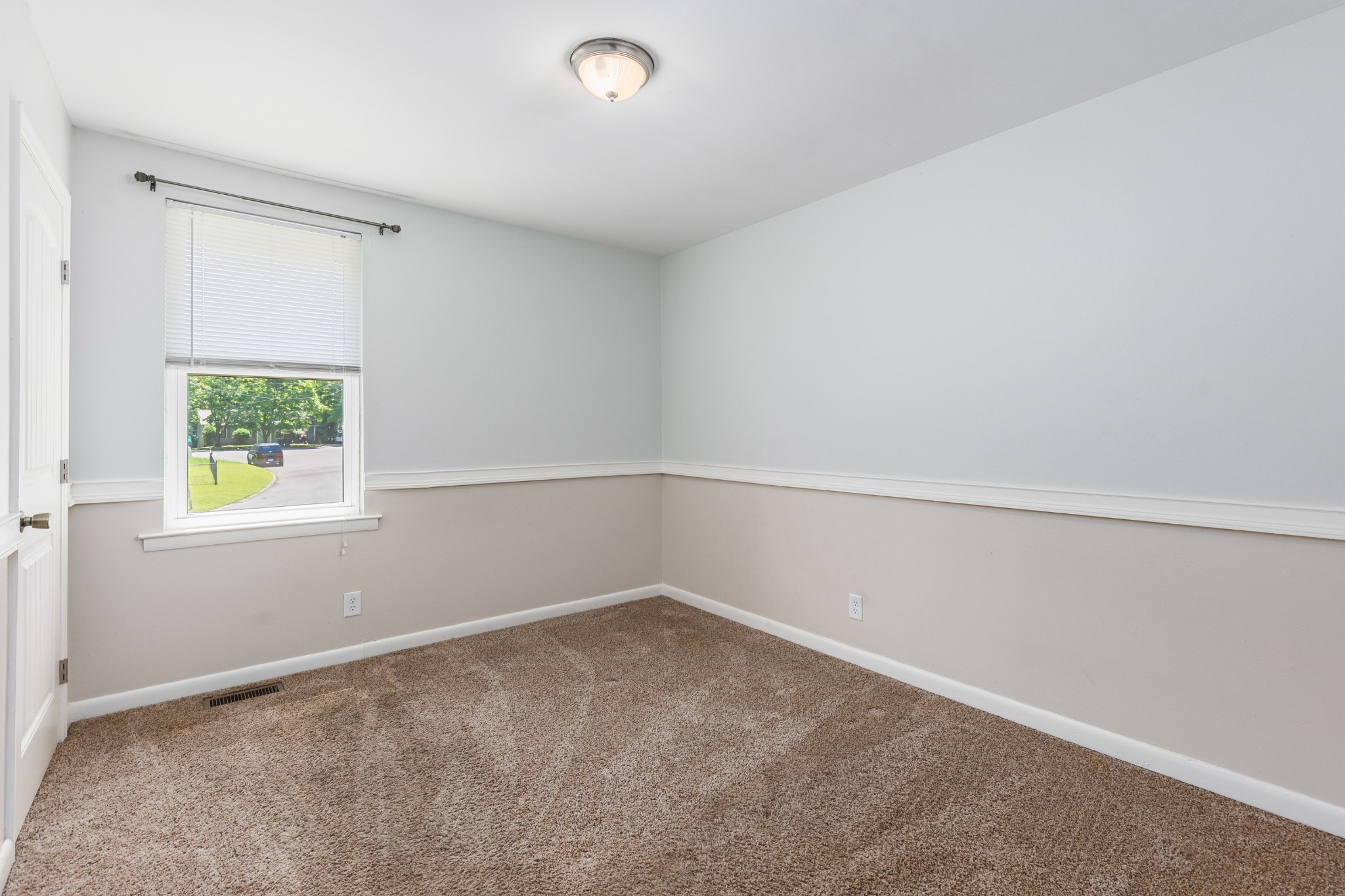 Empty carpeted room with two-tone walls, chair rail, and a window overlooking a leafy street.