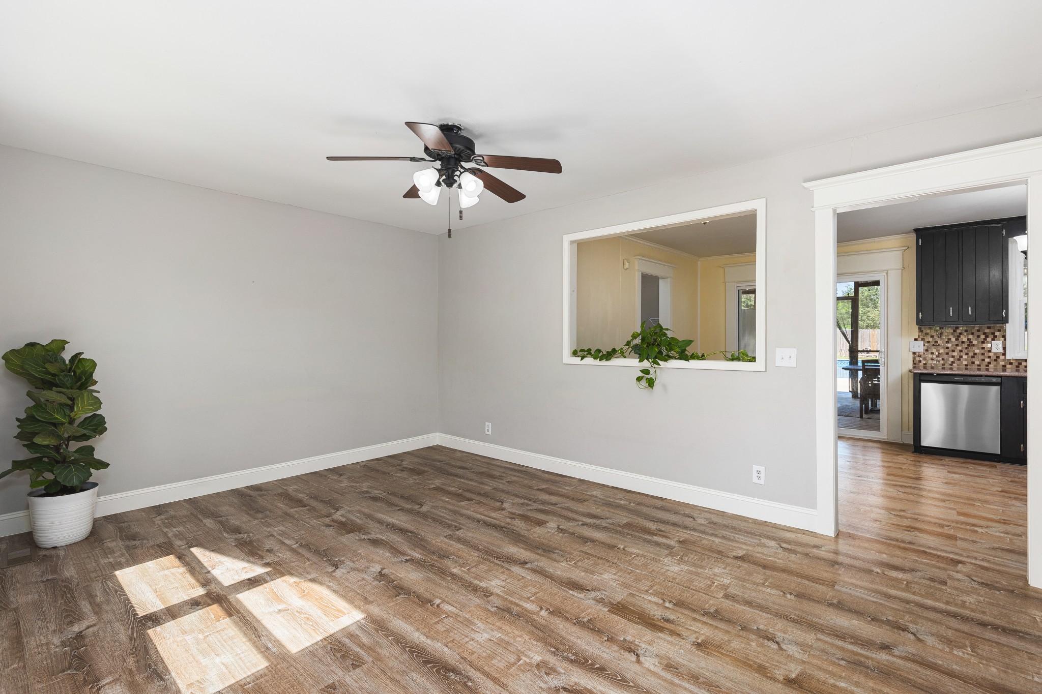 Mostly vacant room with wood-look flooring, a ceiling fan, and an opening to a dark-cabinet kitchen.