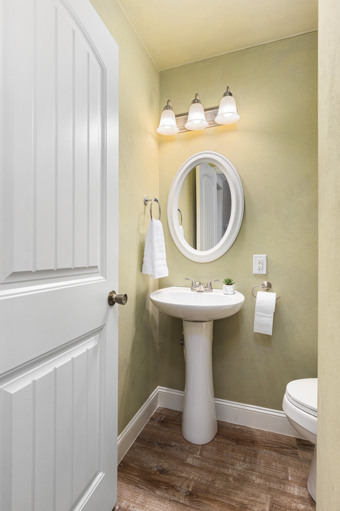 Bathroom with a white pedestal sink, oval mirror, olive-green walls, and dark wood-look flooring.
