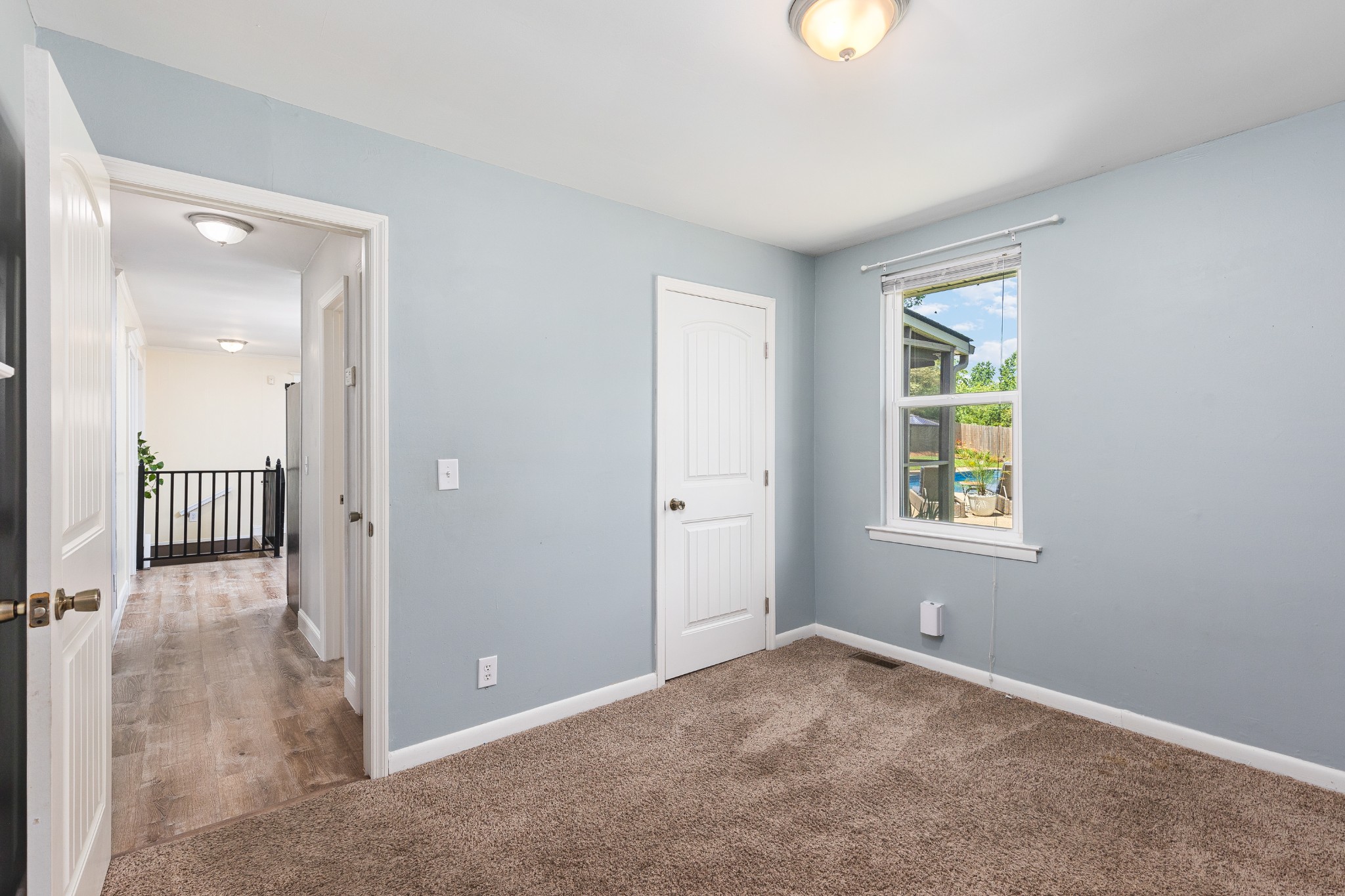 Empty bedroom with pale blue walls, brown carpet, and a window overlooking the backyard pool.