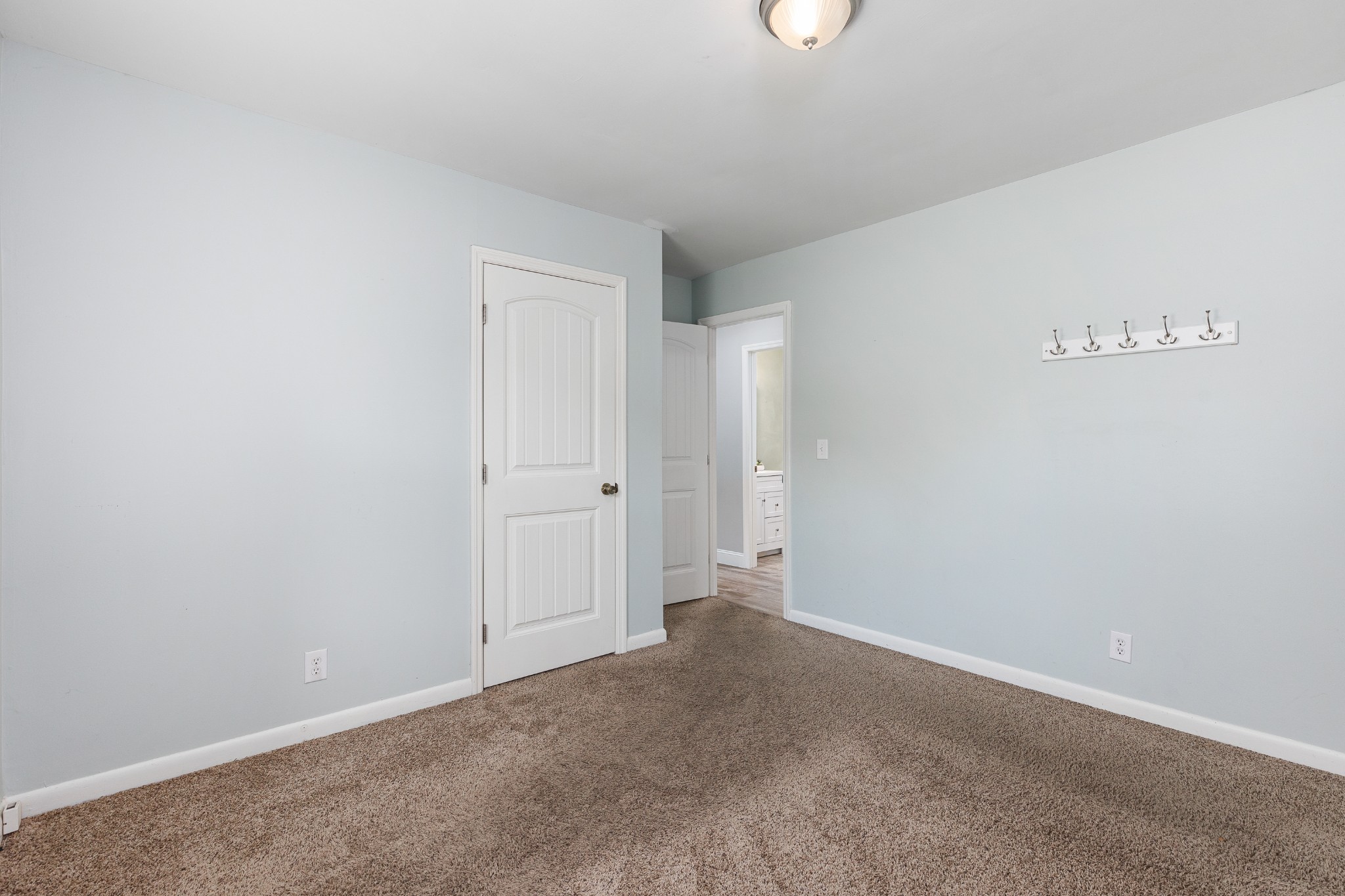 Empty bedroom with brown carpet, pale blue walls, white doors, and wall-mounted coat hooks.