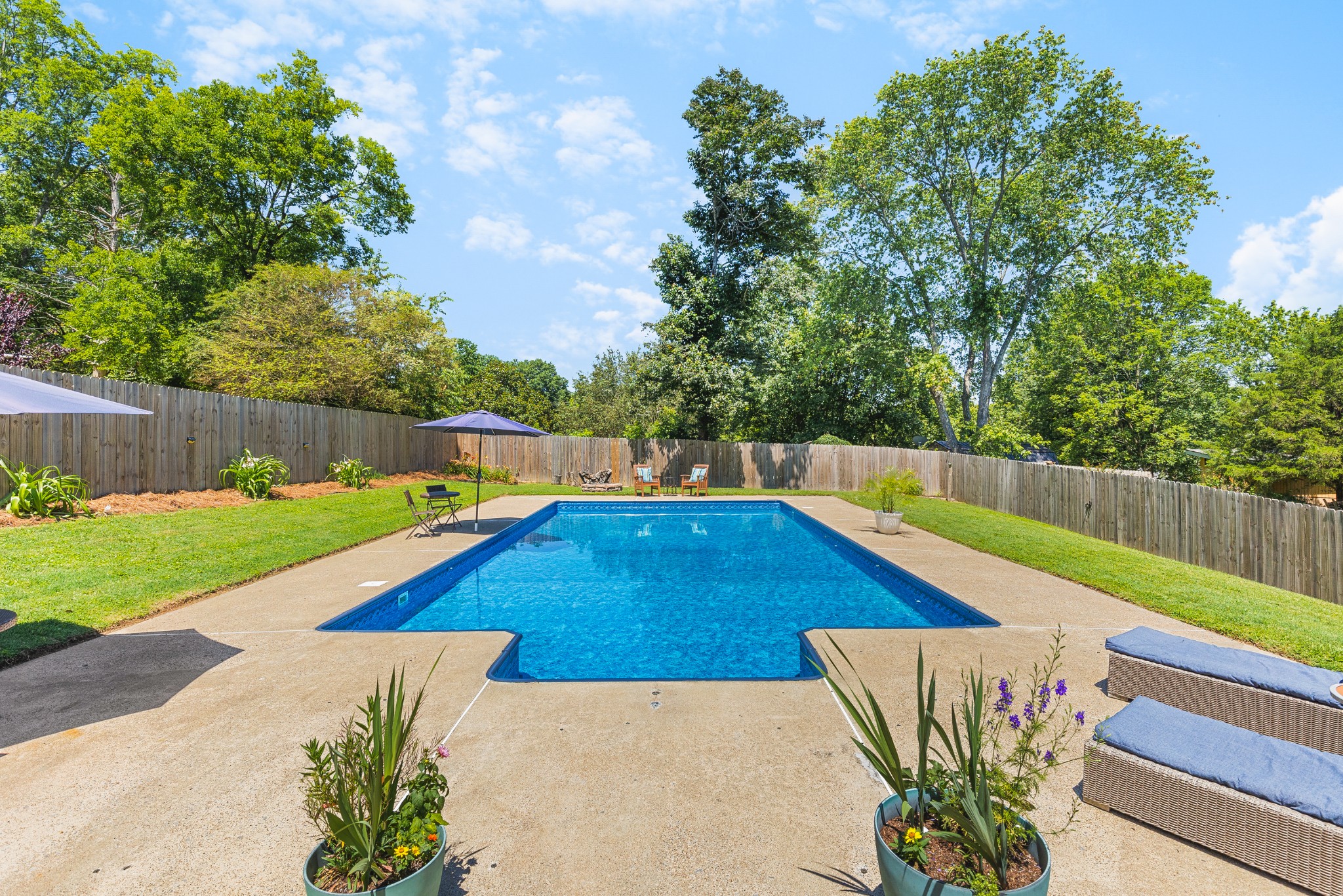 Backyard swimming pool with a concrete patio, umbrellas, and a fenced, tree-lined lawn.