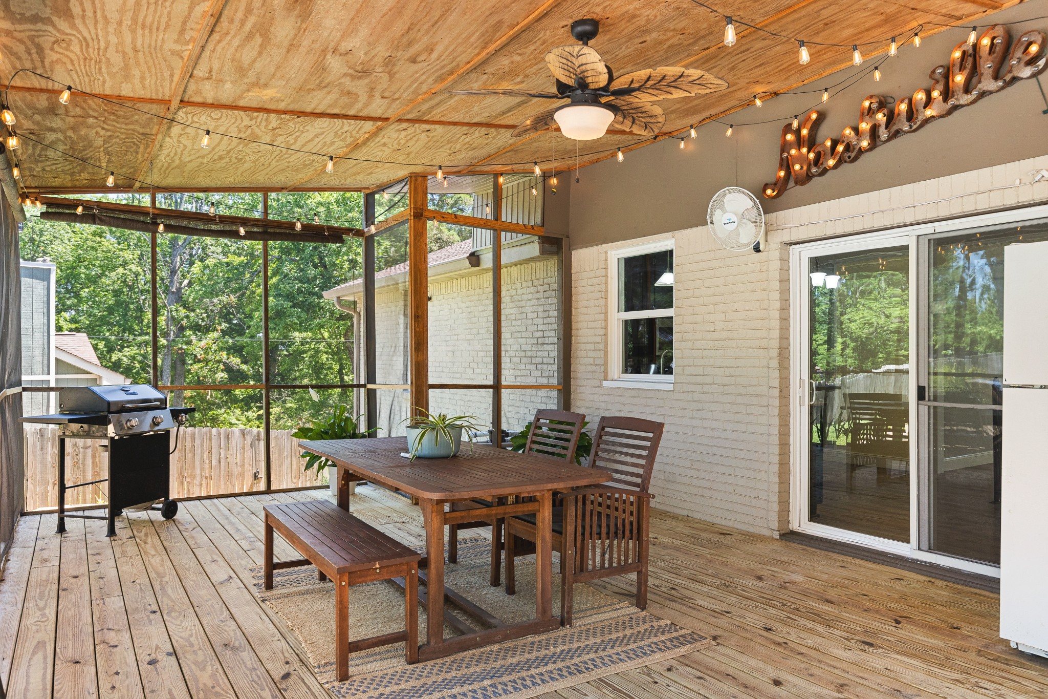 Screened porch with wooden dining set, string lights, ceiling fan, and barbecue grill.