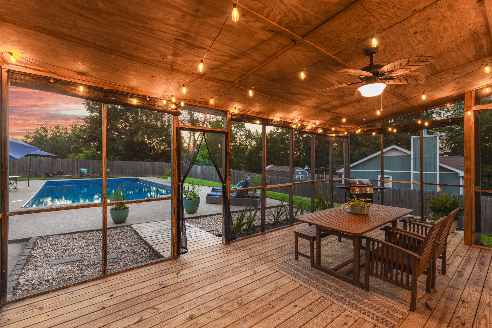 Screened wooden deck with string lights and dining furniture overlooking a backyard pool at sunset.