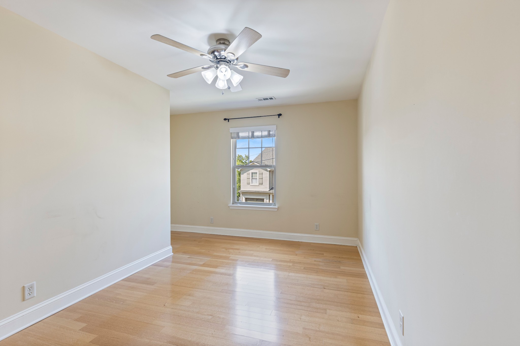 Empty bedroom with hardwood floors, ceiling fan, and a window overlooking a neighboring house.