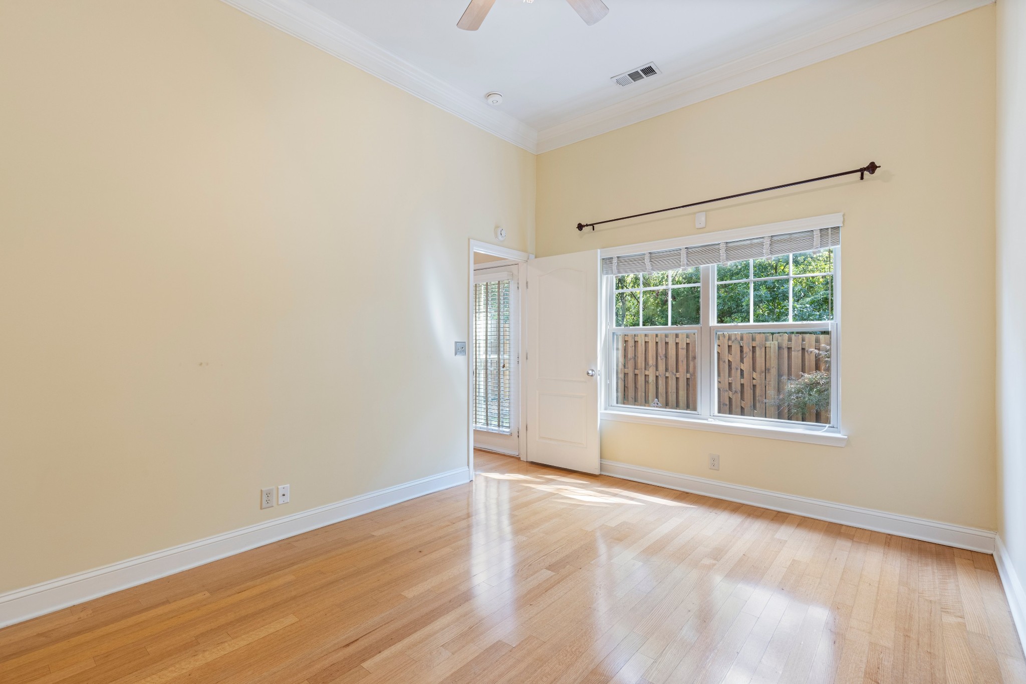 Empty bedroom with hardwood floors and a large window overlooking a wooden fence and greenery.