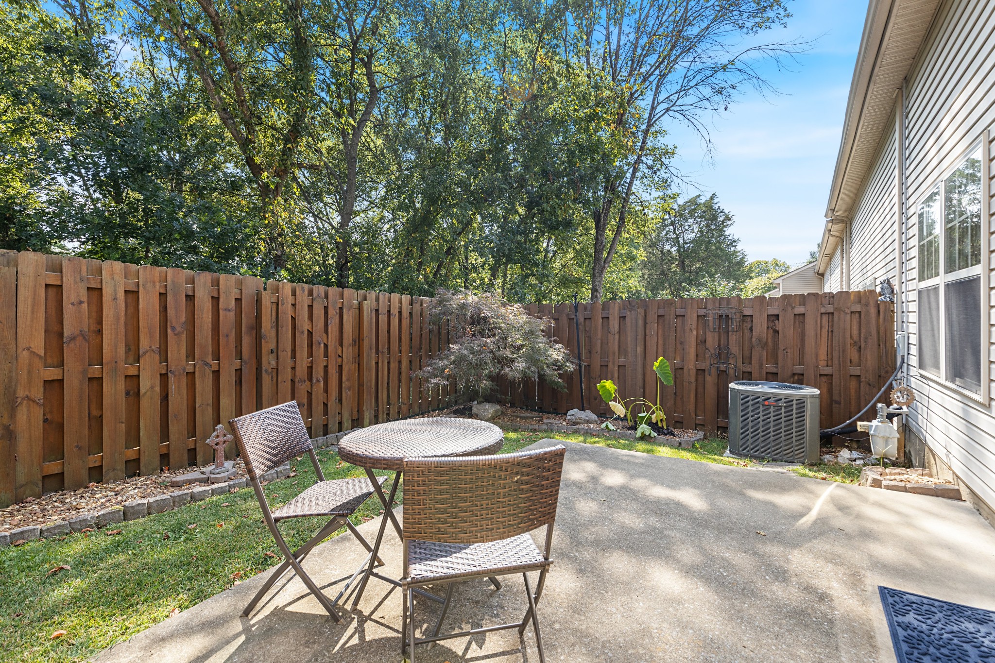 Fenced backyard patio with wicker seating, mature trees, and a landscaped border beside the home.