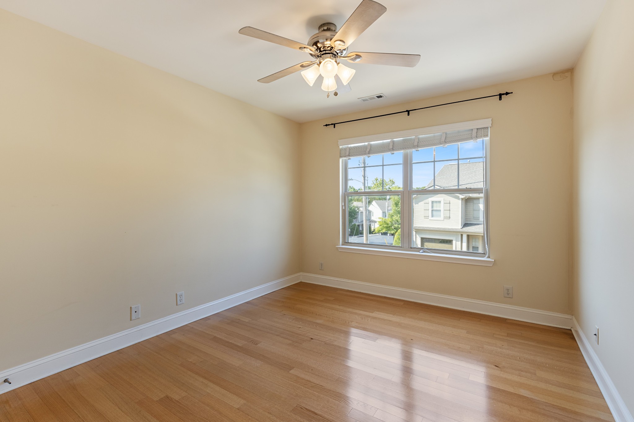 Empty bedroom with hardwood floors, ceiling fan, and a large window overlooking neighboring homes.