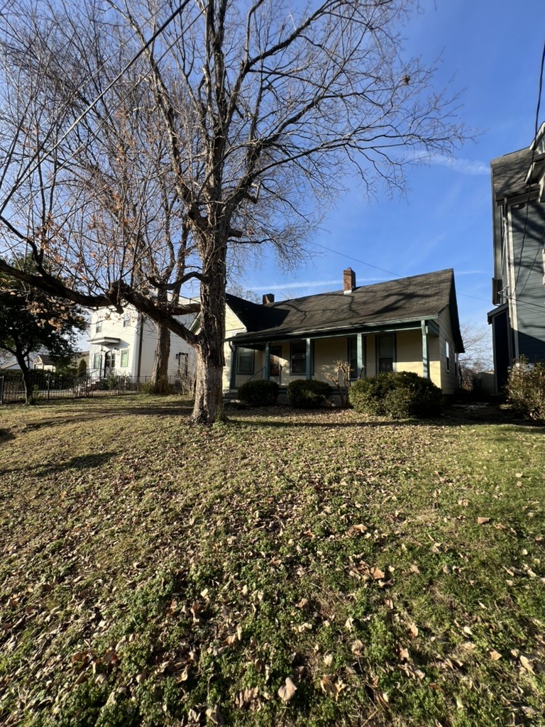 Single-story house with a covered porch behind a large leafless tree and leaf-strewn lawn.