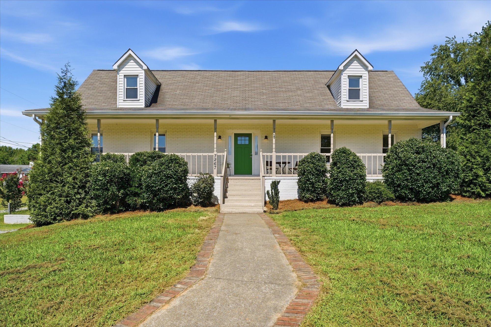 Light-colored house with a covered porch, green door, roof dormers, and brick-edged walkway.