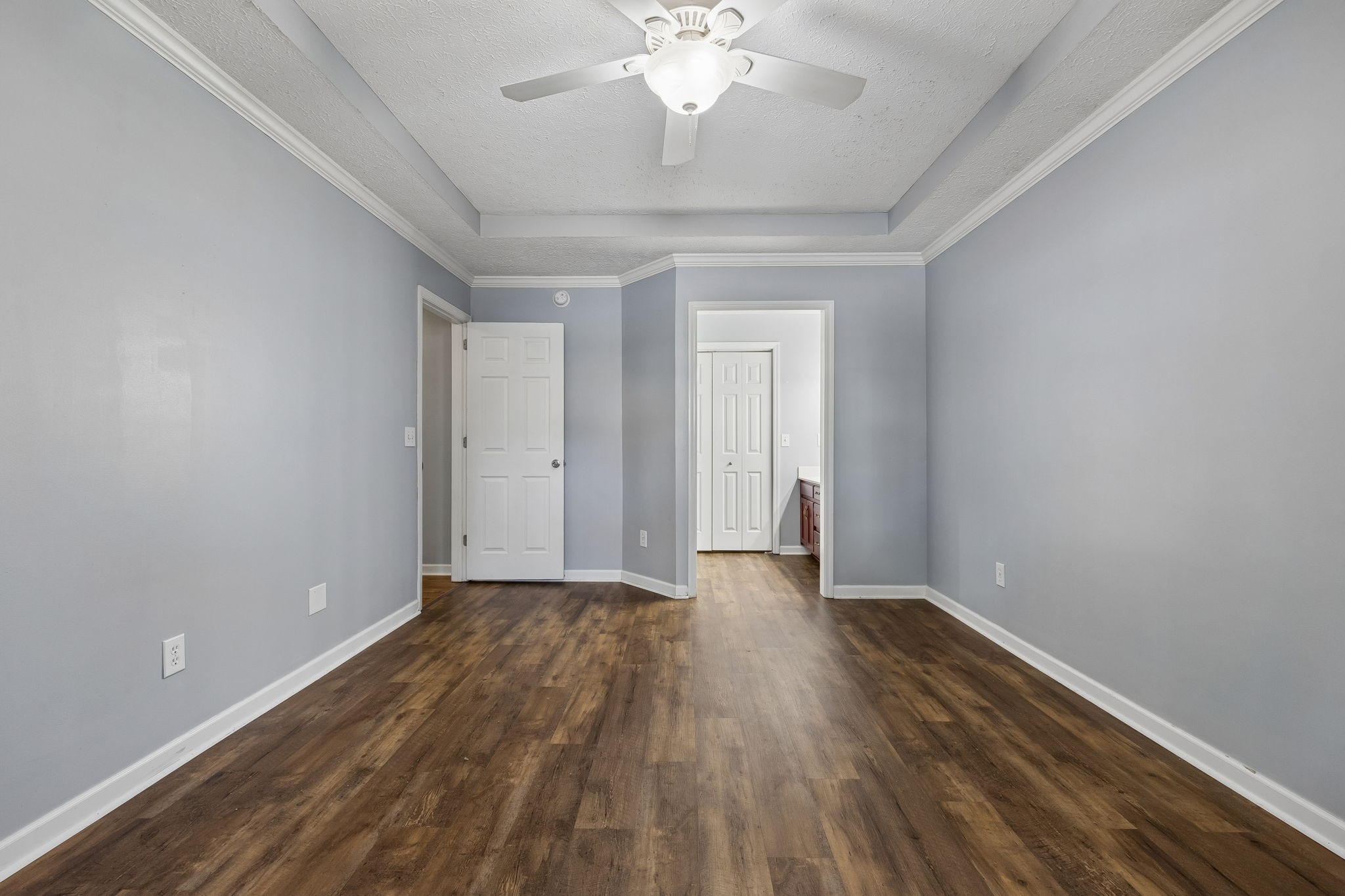 Empty gray-walled room with dark wood-look flooring, tray ceiling, and ceiling fan.