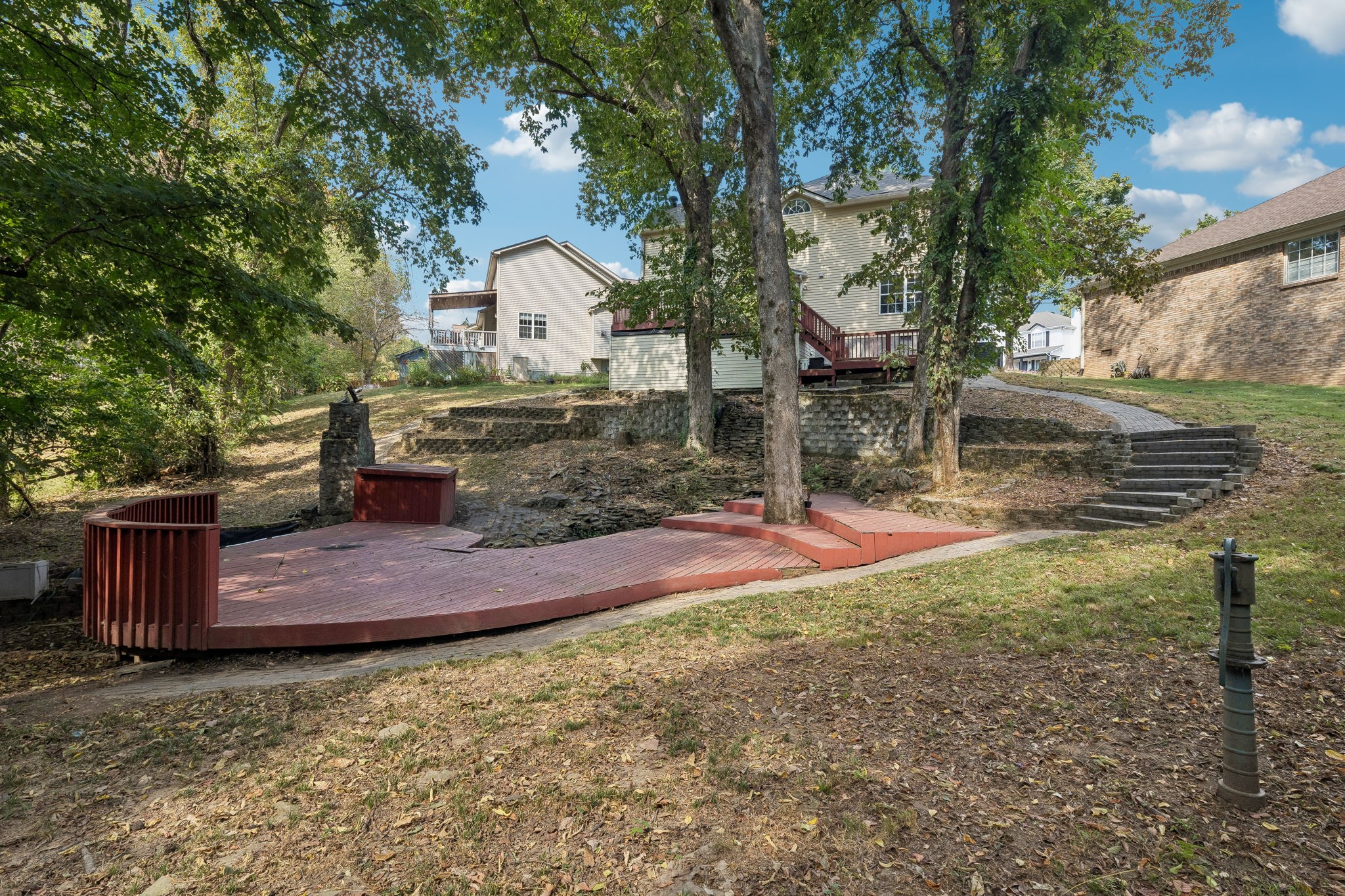 Backyard deck curves around mature trees, with stone retaining walls and steps leading up to the house.