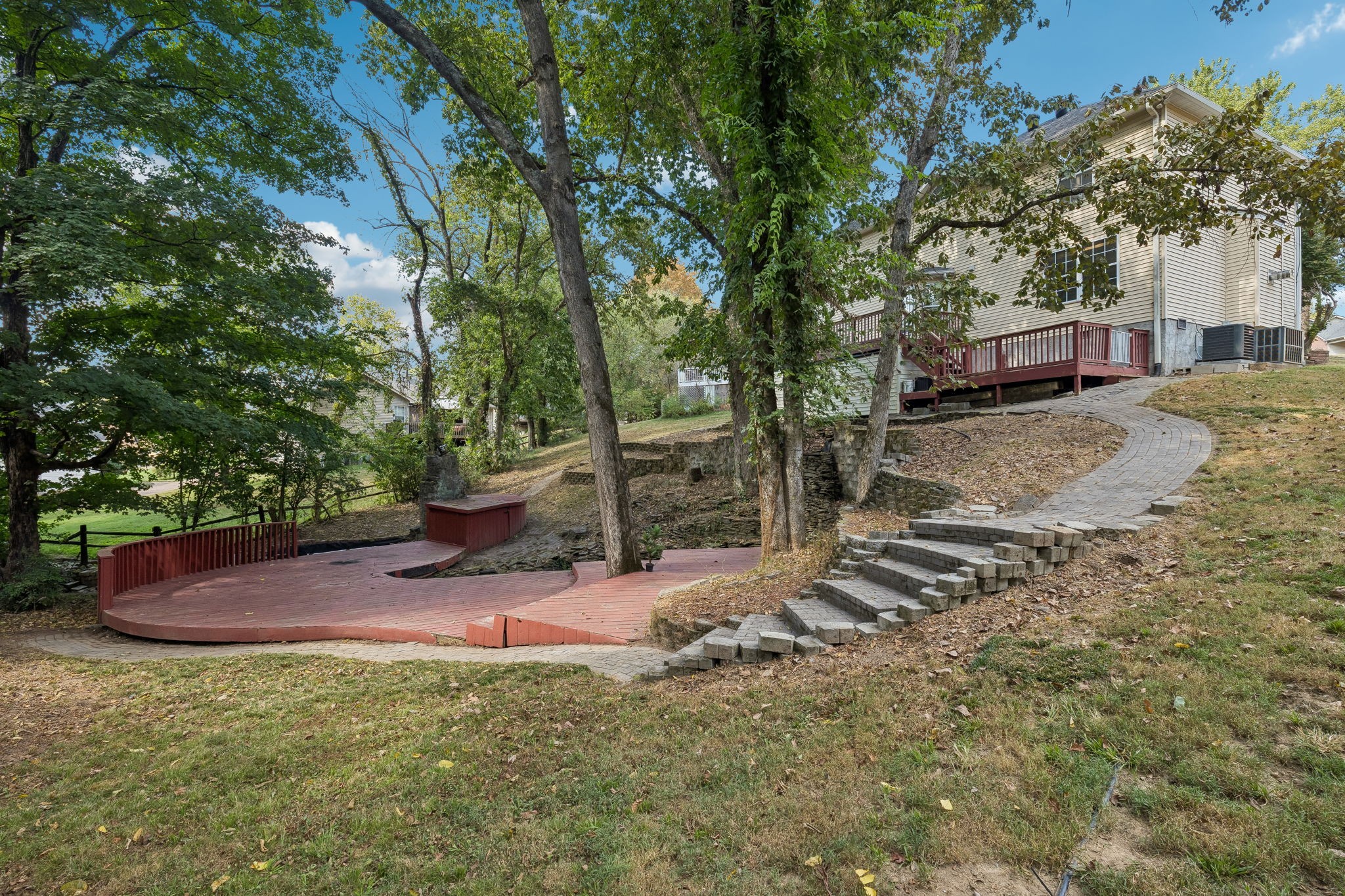 House rear with raised red deck, lower deck, and curved stone steps through a wooded yard.