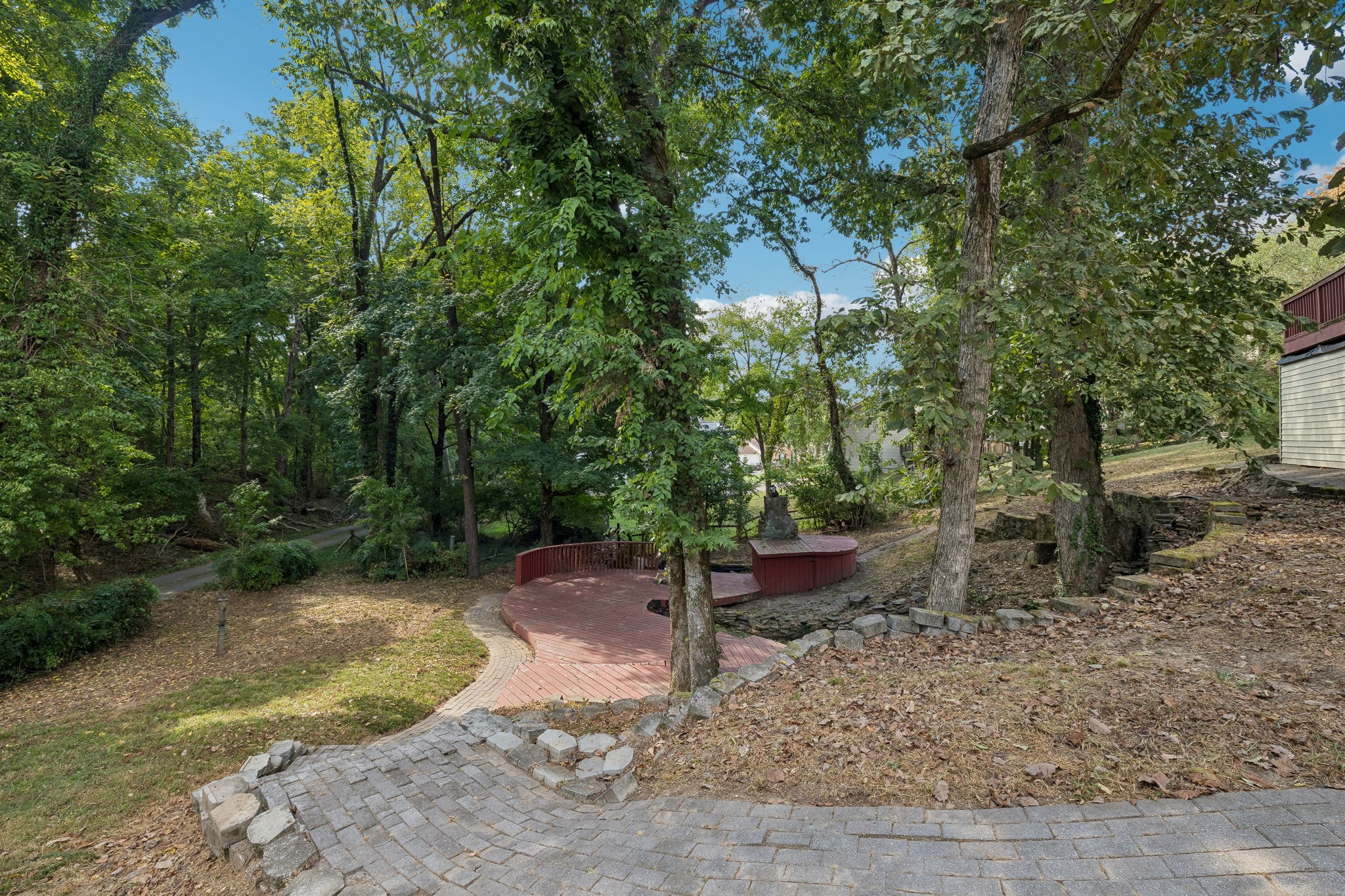 Wooded backyard with a tiered red deck, curved paver path, and stone retaining walls.