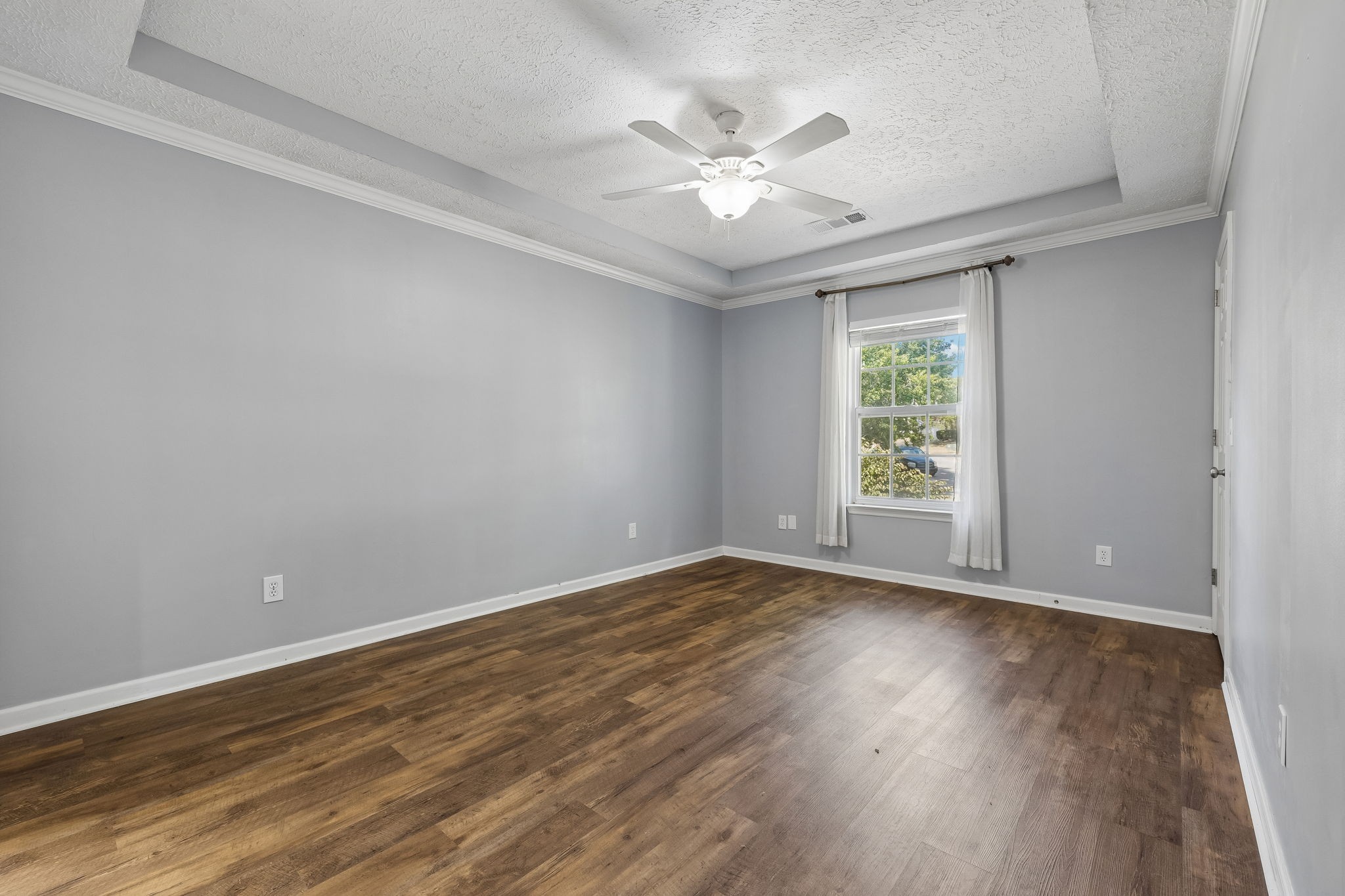 Empty room with brown wood-look floors, gray walls, a tray ceiling, and a curtained window.