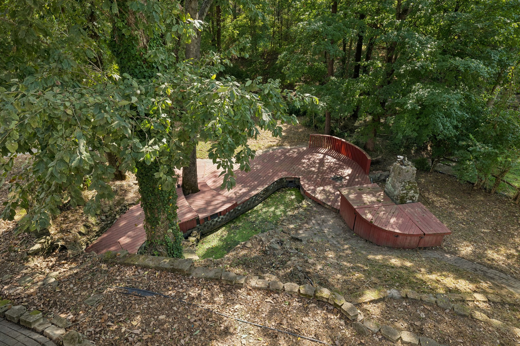 Curved wooden deck with red railings nestled among mature trees in a wooded backyard.